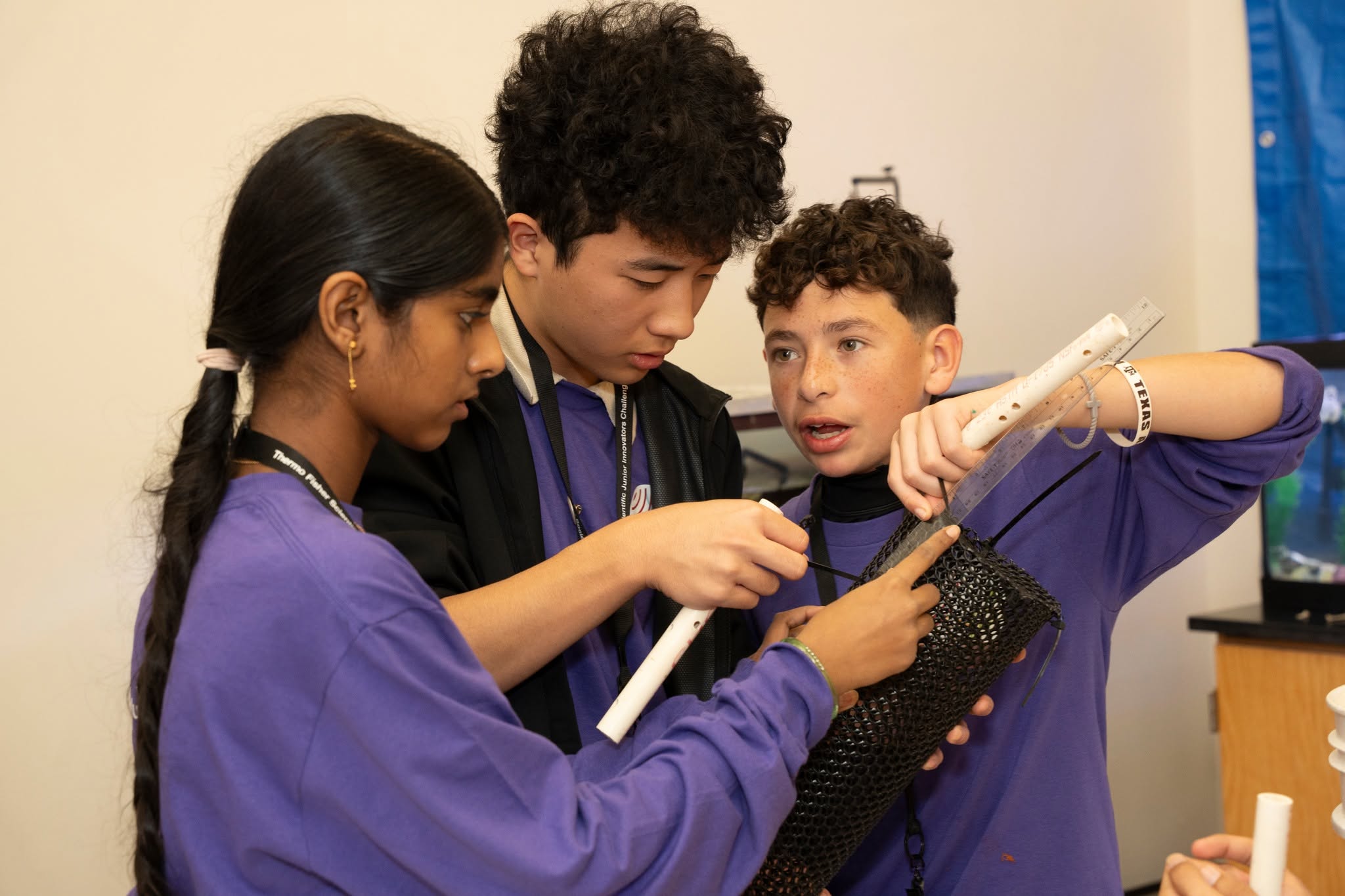 Three students collaborate on a science project, measuring a black mesh cylinder with white pipes and a ruler.