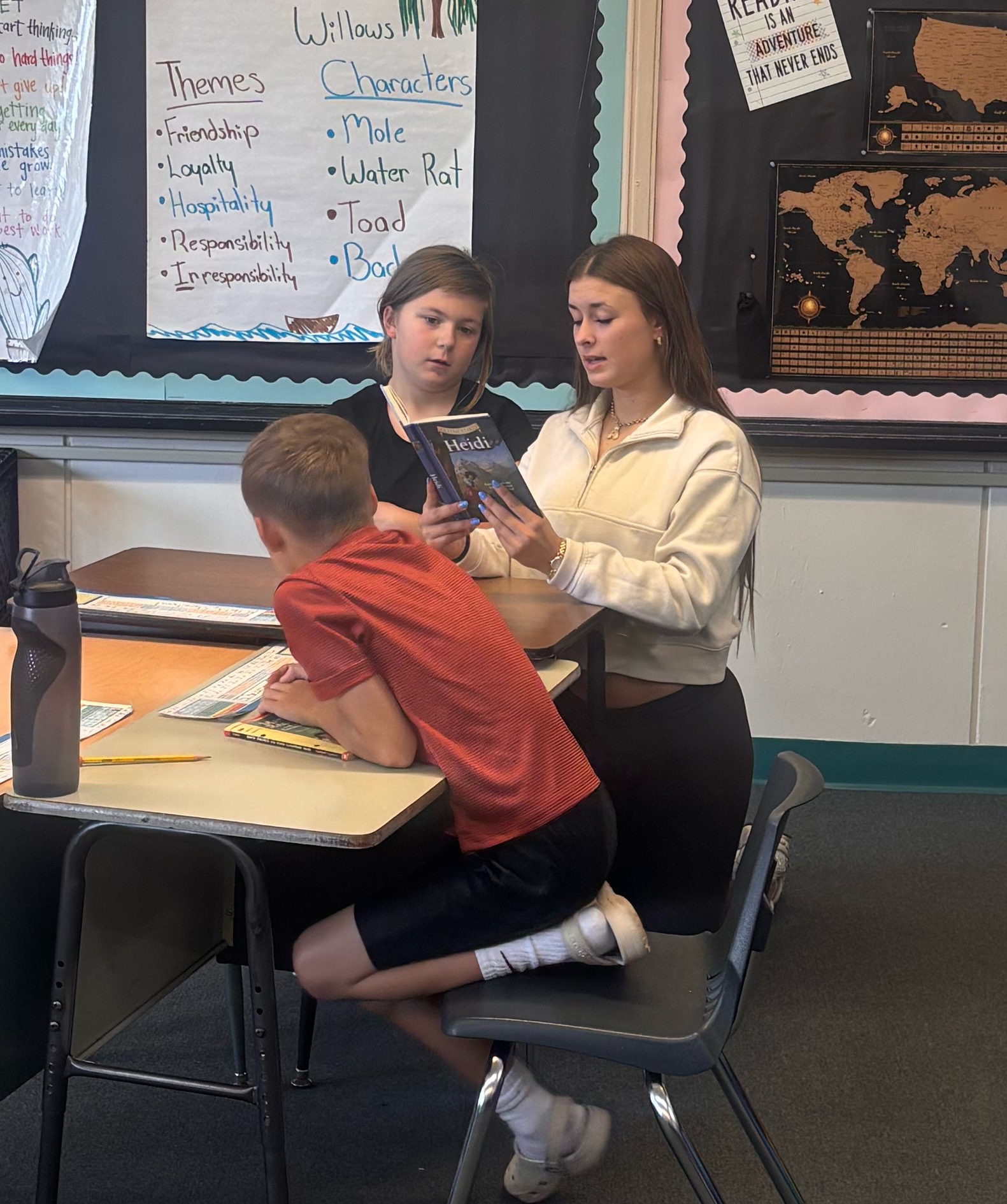 A teacher assists a student reading a Heidi book in a classroom with posters on the wall.