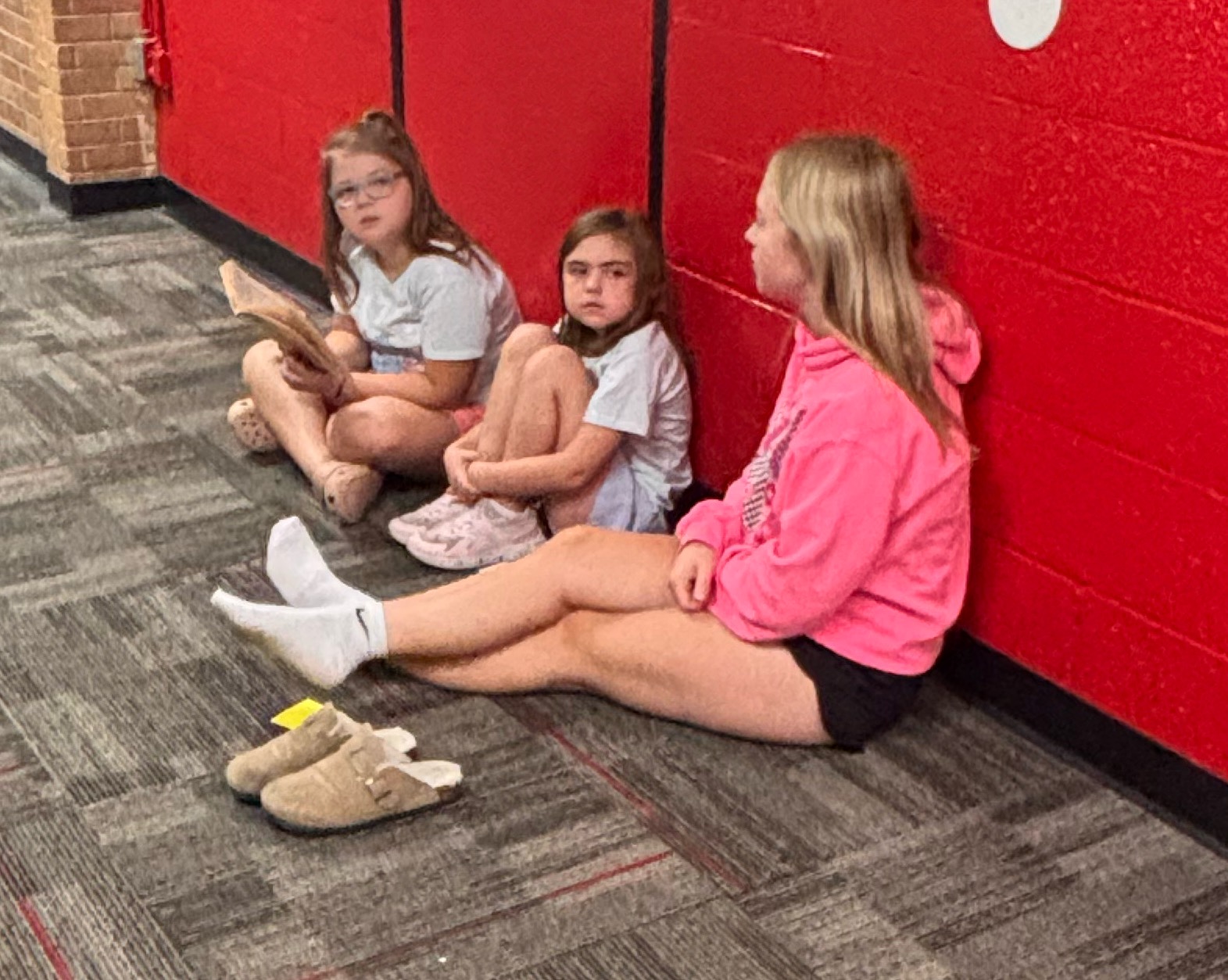 Three young people sit on the gray carpeted floor, leaning against a bright red wall in a hallway.