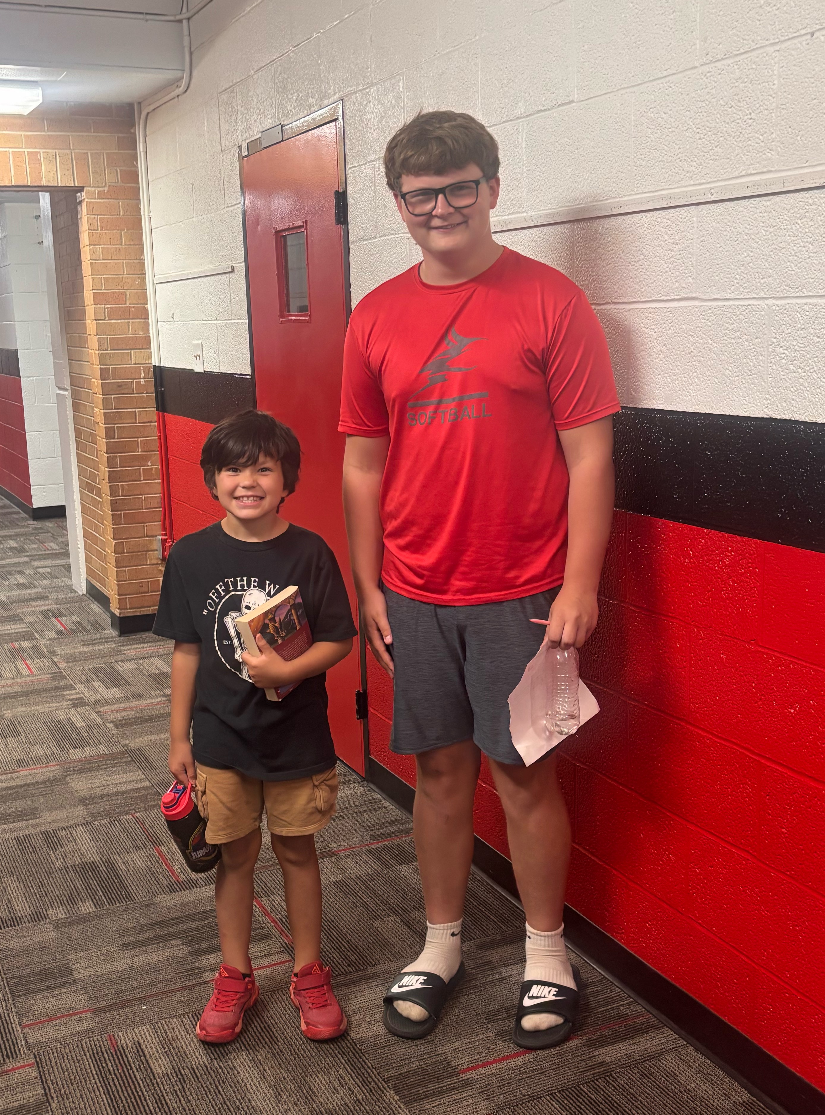 Two boys stand smiling together in a school hallway with red and black block walls.