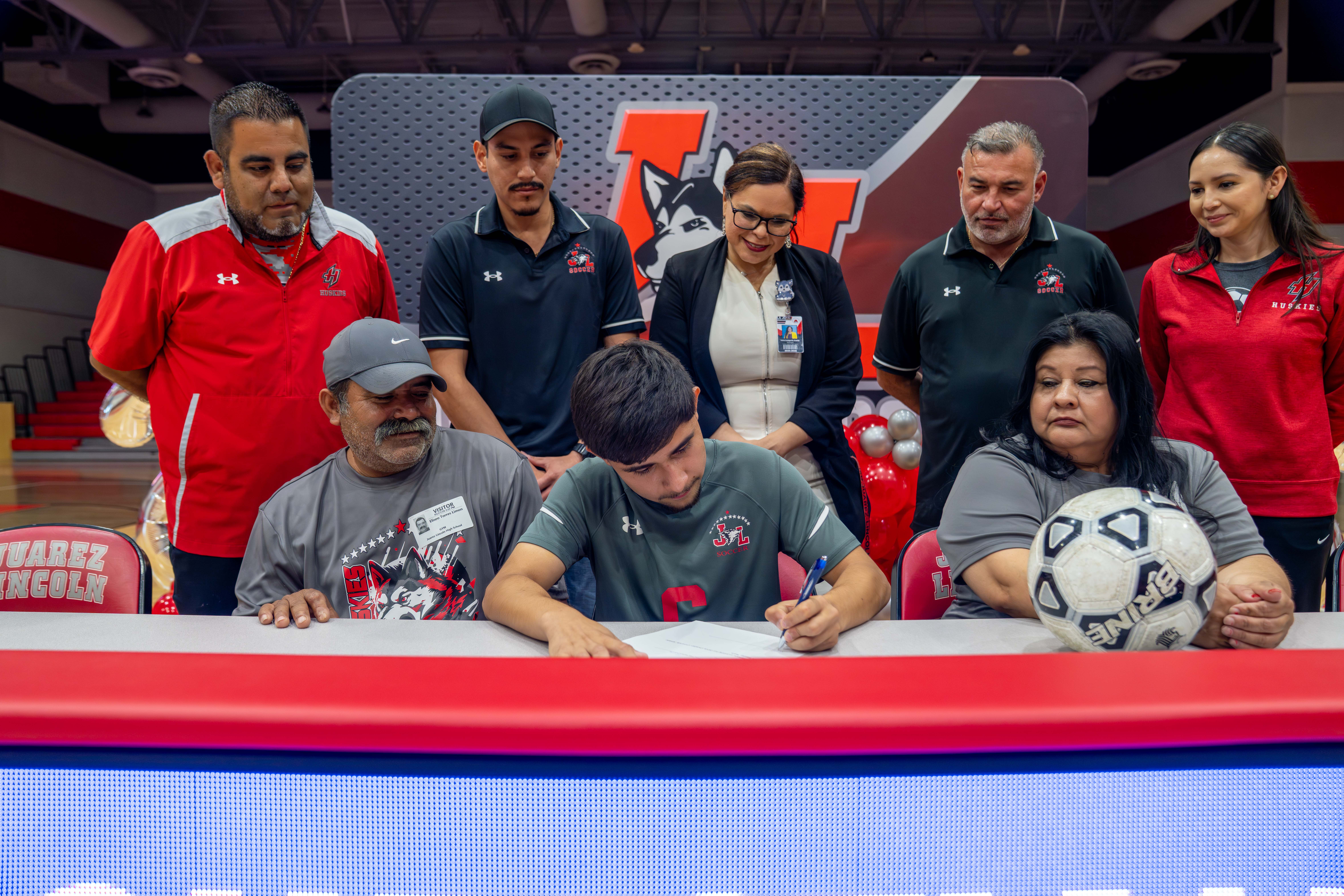 A young soccer player signs a document at a table, surrounded by family and coaches.