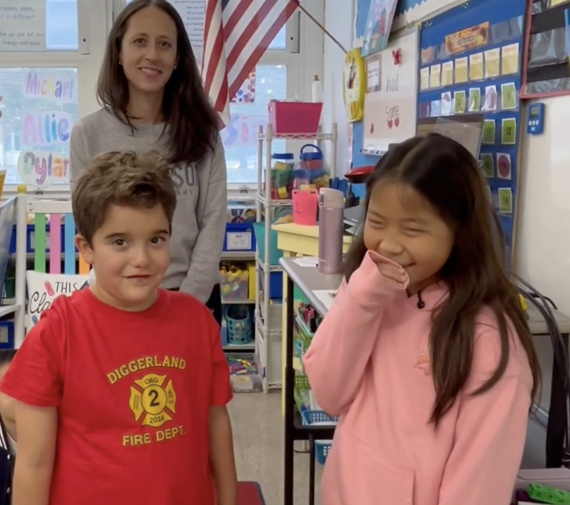 A teacher stands behind two students smiling in a brightly decorated elementary classroom.