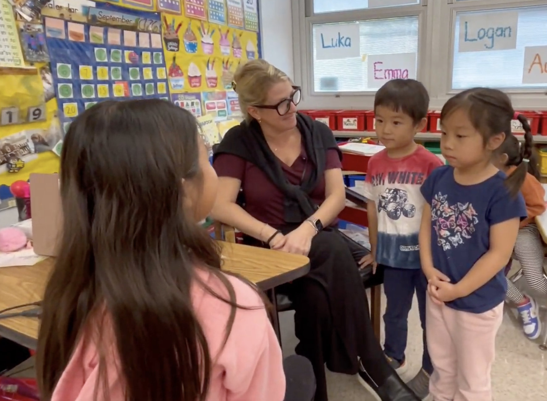 A teacher sits in a classroom while students gather around to talk and learn.