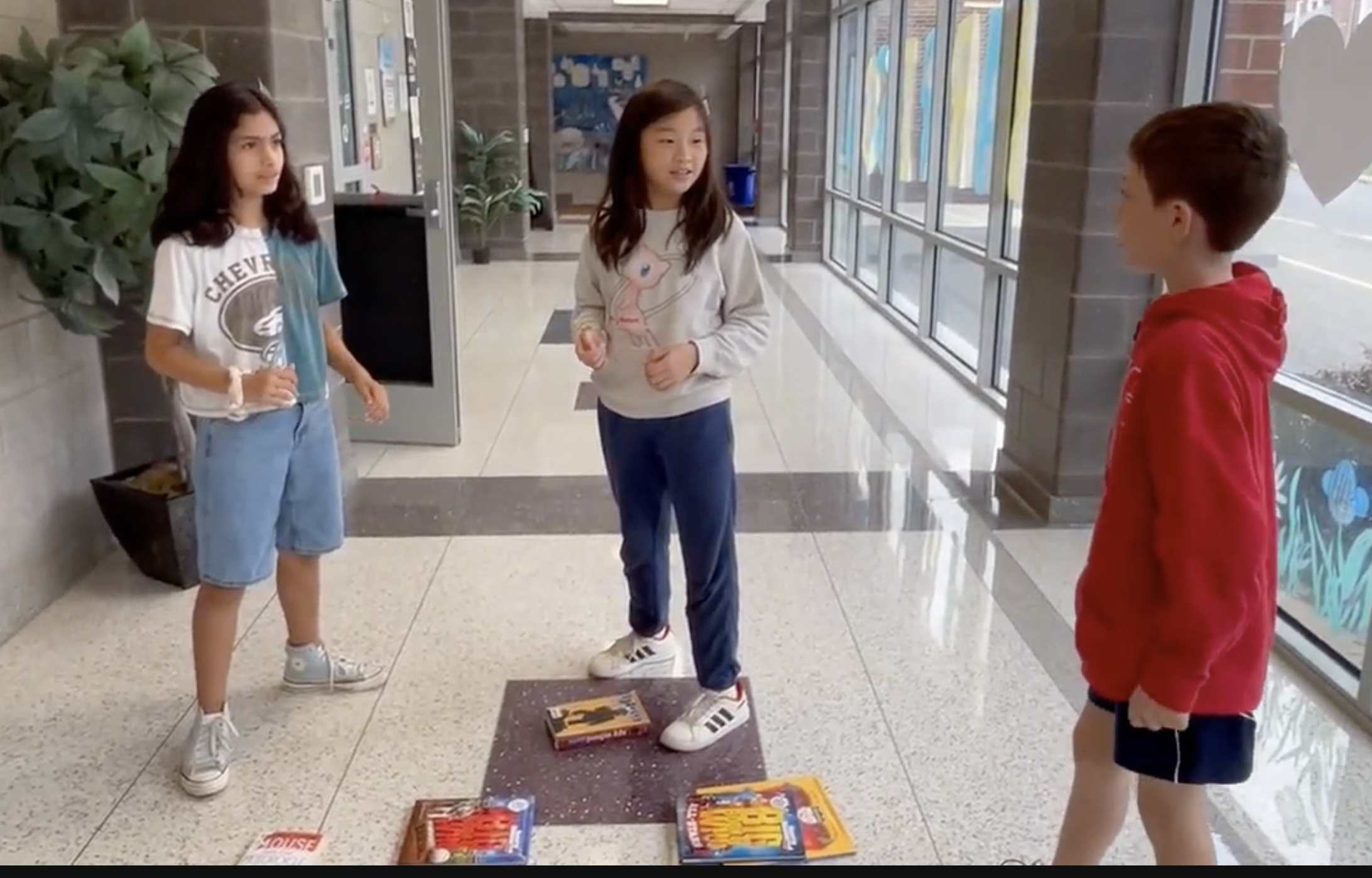 Three children stand in a school hallway with books arranged on the floor.