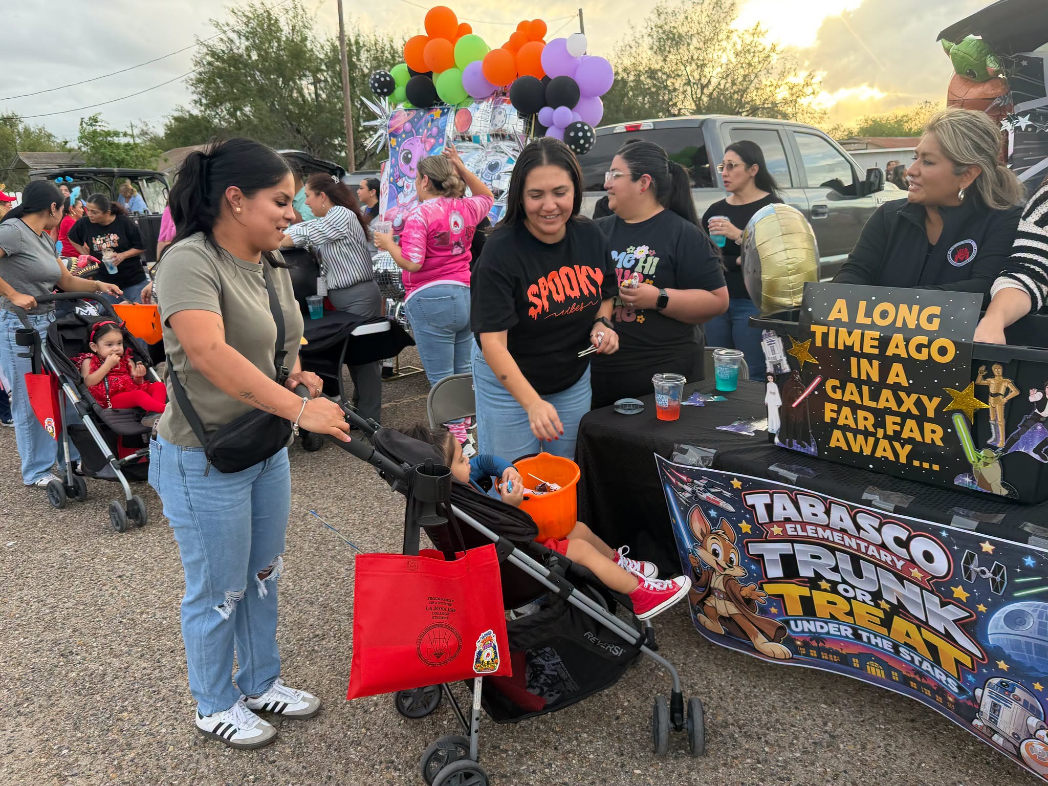 People gather at an outdoor trunk-or-treat event with colorful balloons and decorated car setups.