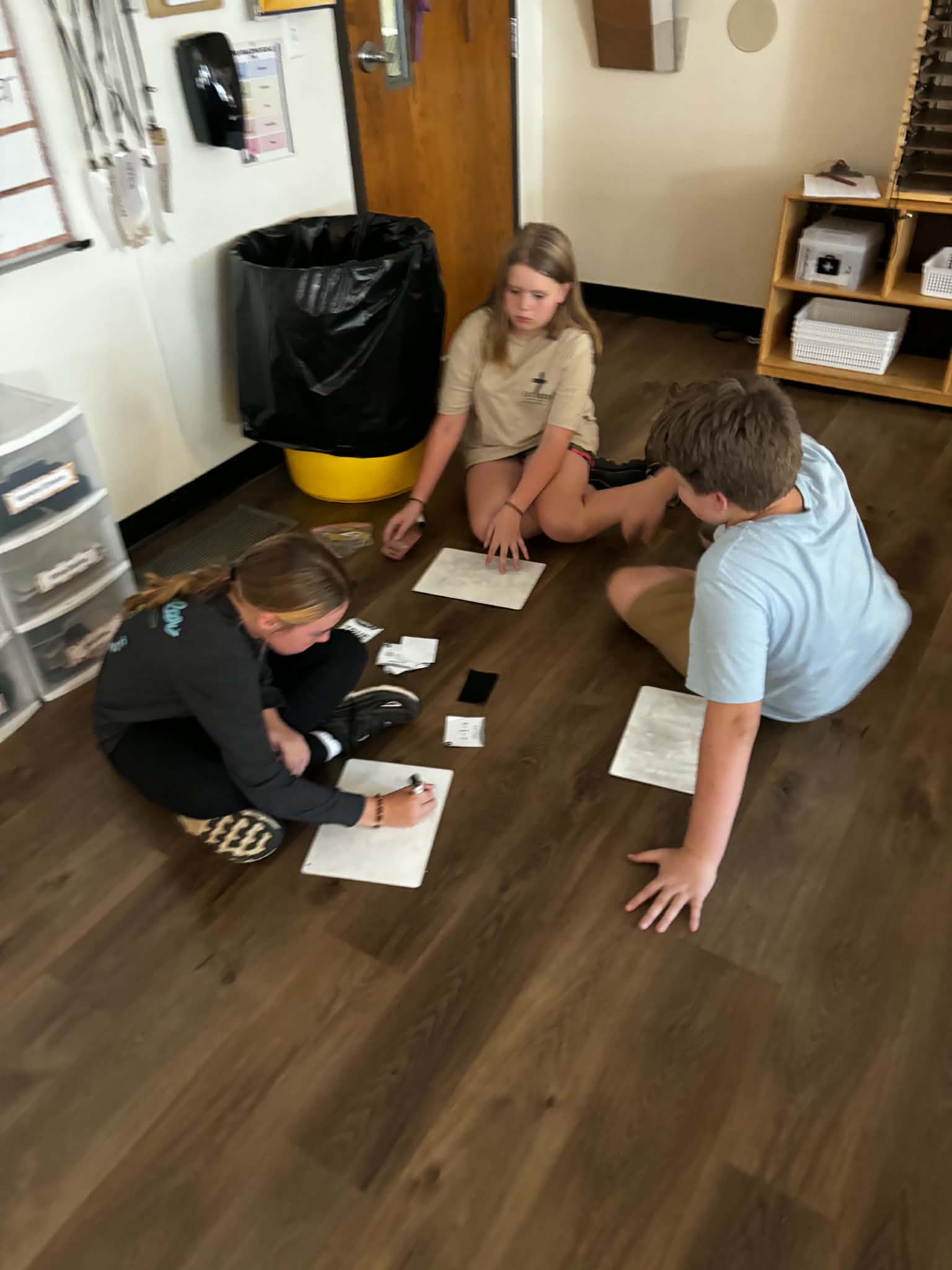 Three students sit on a classroom floor, working together around small whiteboards and cards.