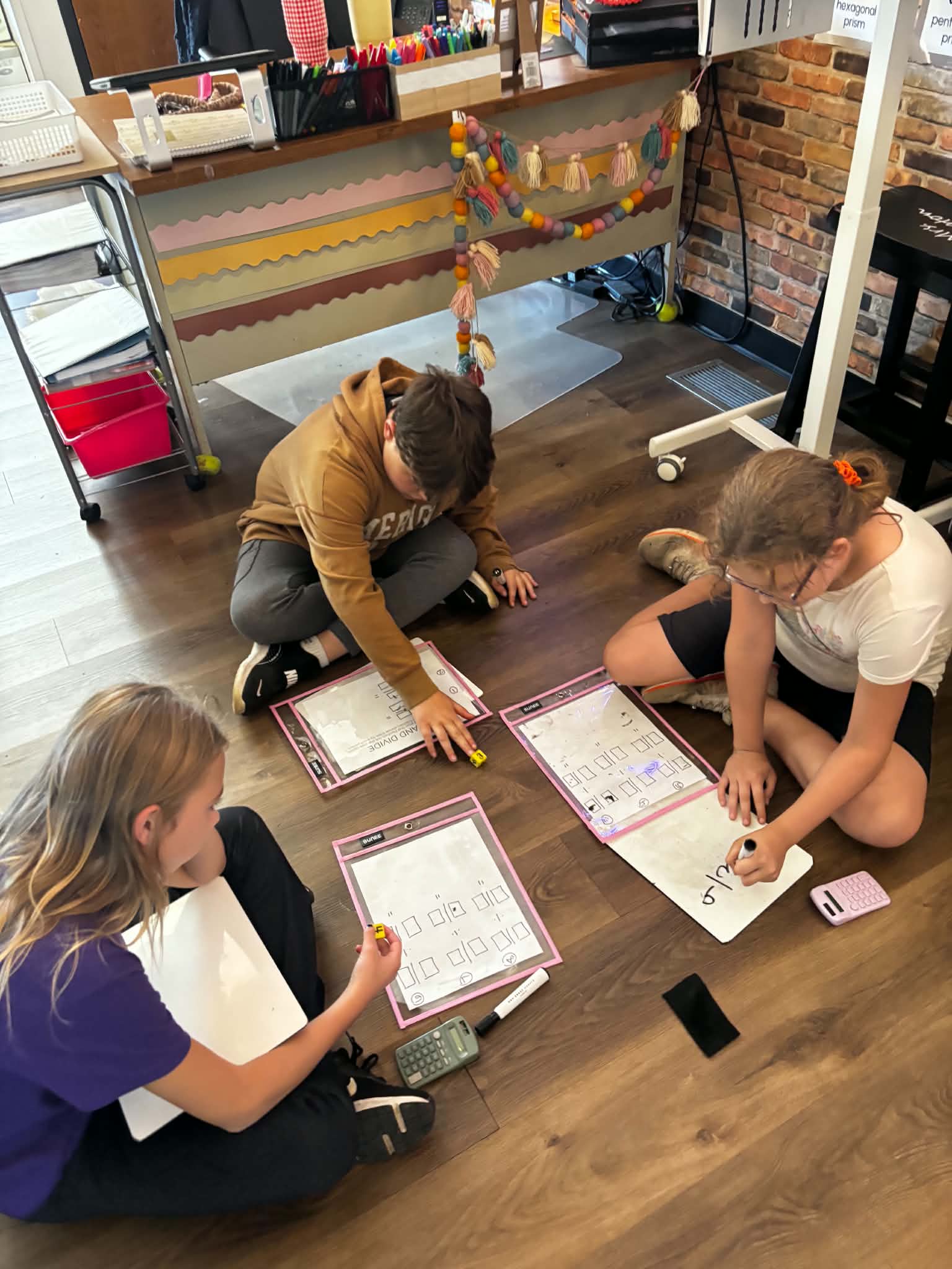 Three students sit on the classroom floor, working on math worksheets and whiteboards.