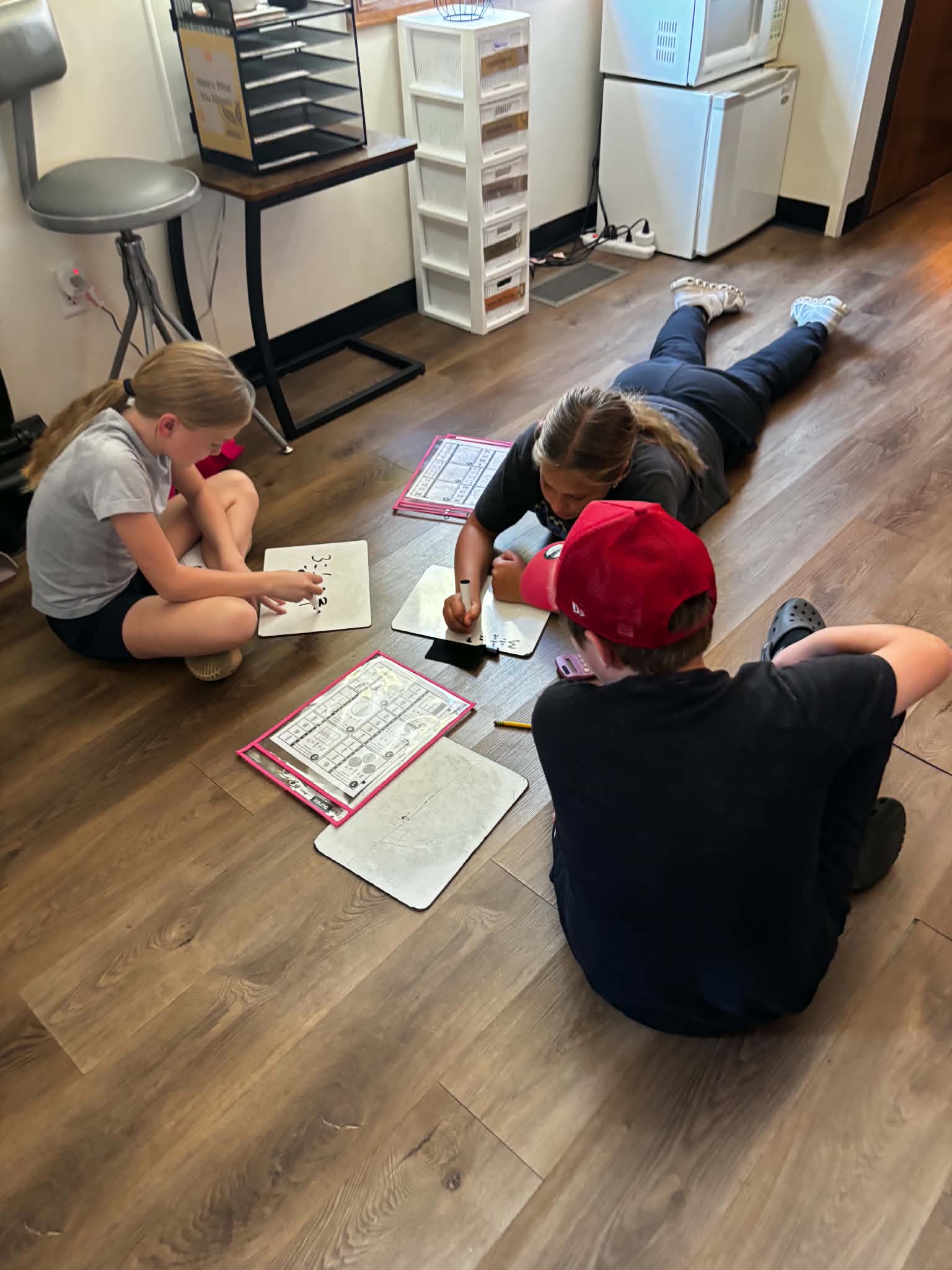 Three children sit and lie on a wooden floor, working on math worksheets with dry-erase boards.