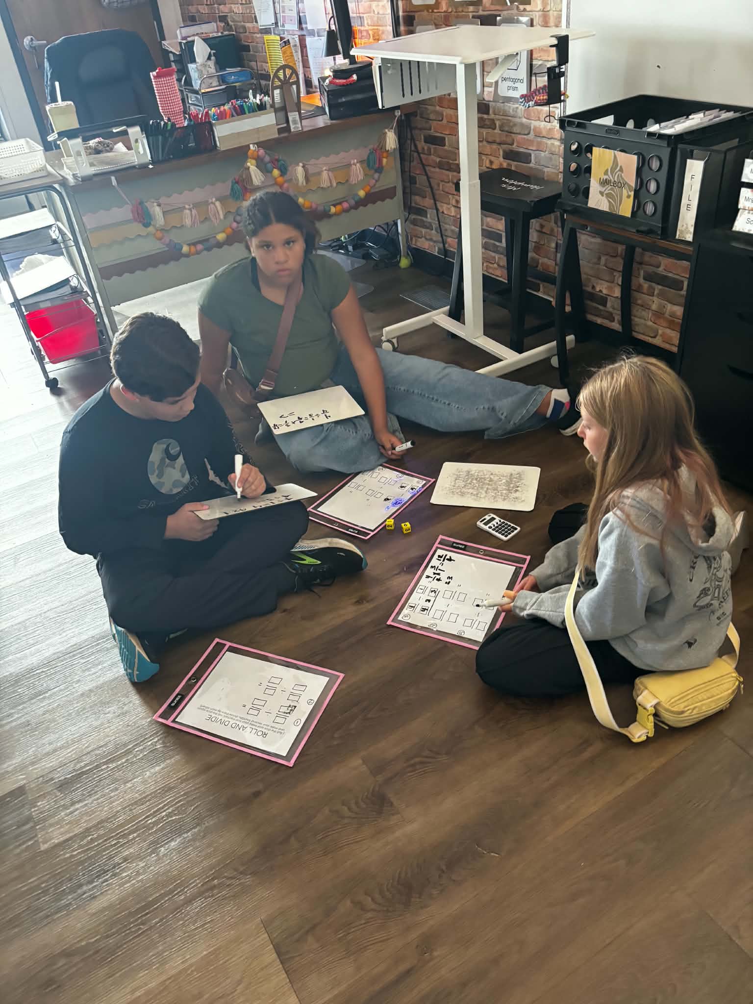 Three students sit on a classroom floor, working on math worksheets with dry-erase markers.
