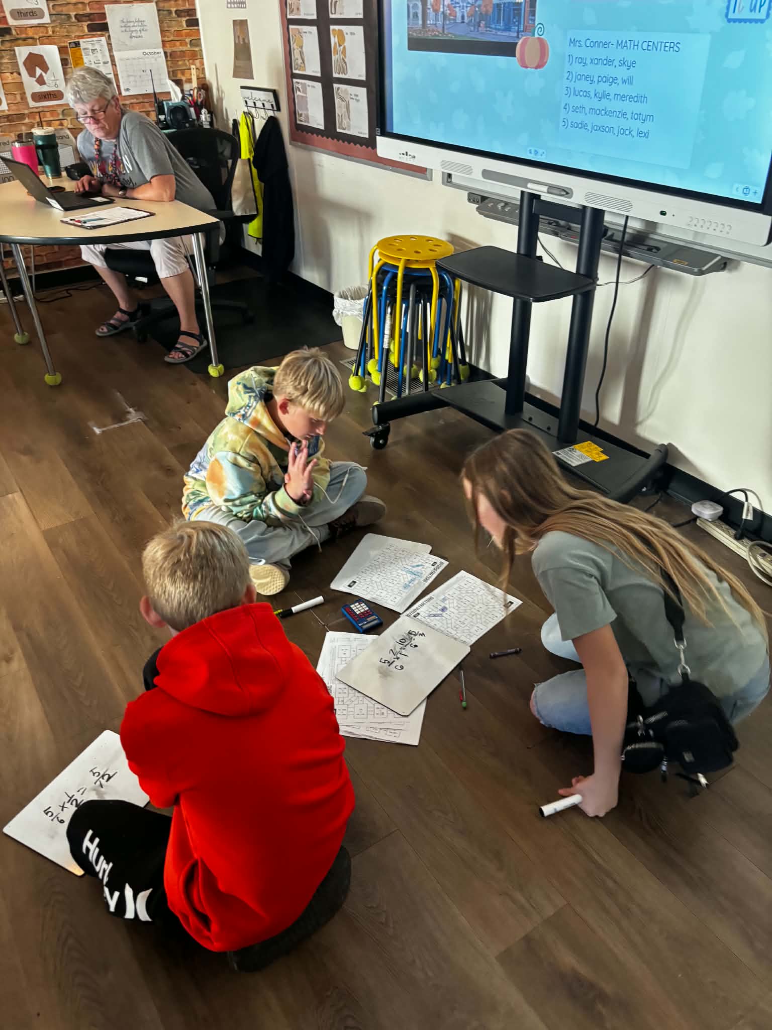 Three students work together on math assignments spread across a classroom floor.