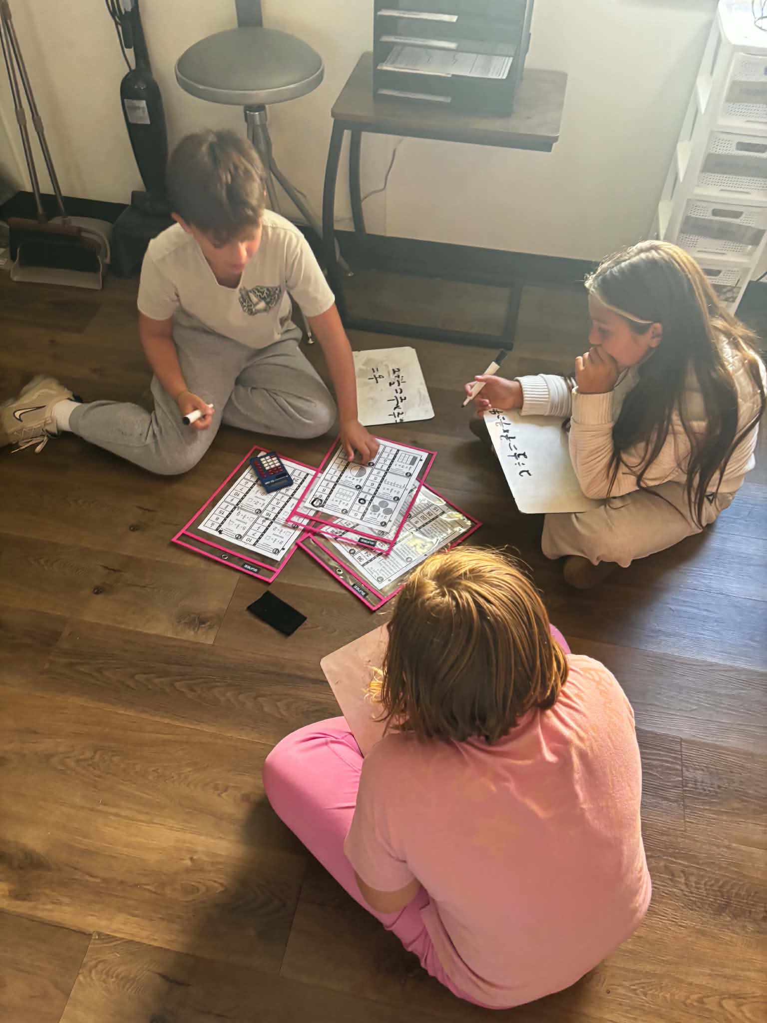 Three children sit on a hardwood floor, working collaboratively on math worksheets and writing on small whiteboards.