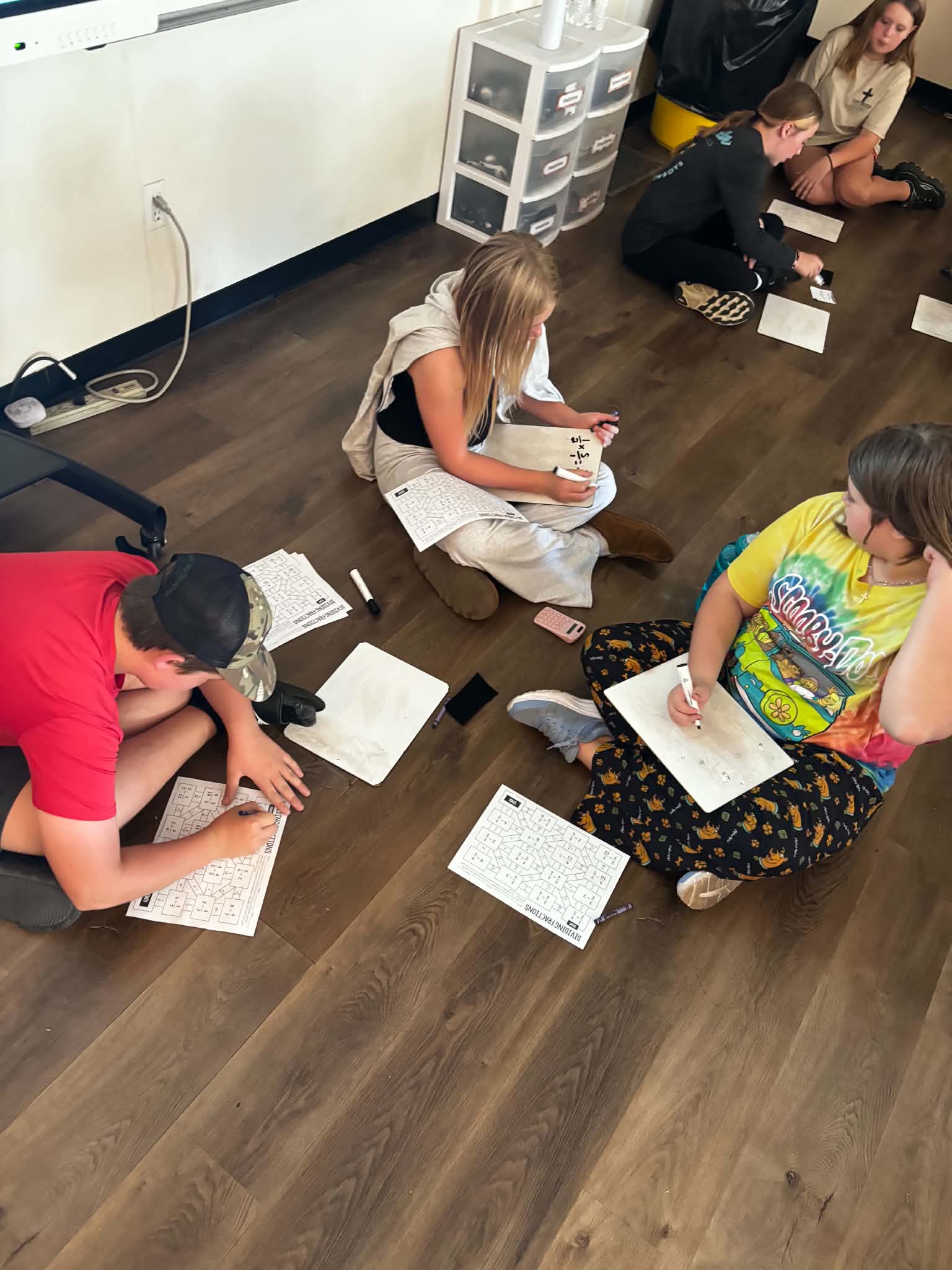 Students sit on a classroom floor, working on math worksheets and writing on small whiteboards.