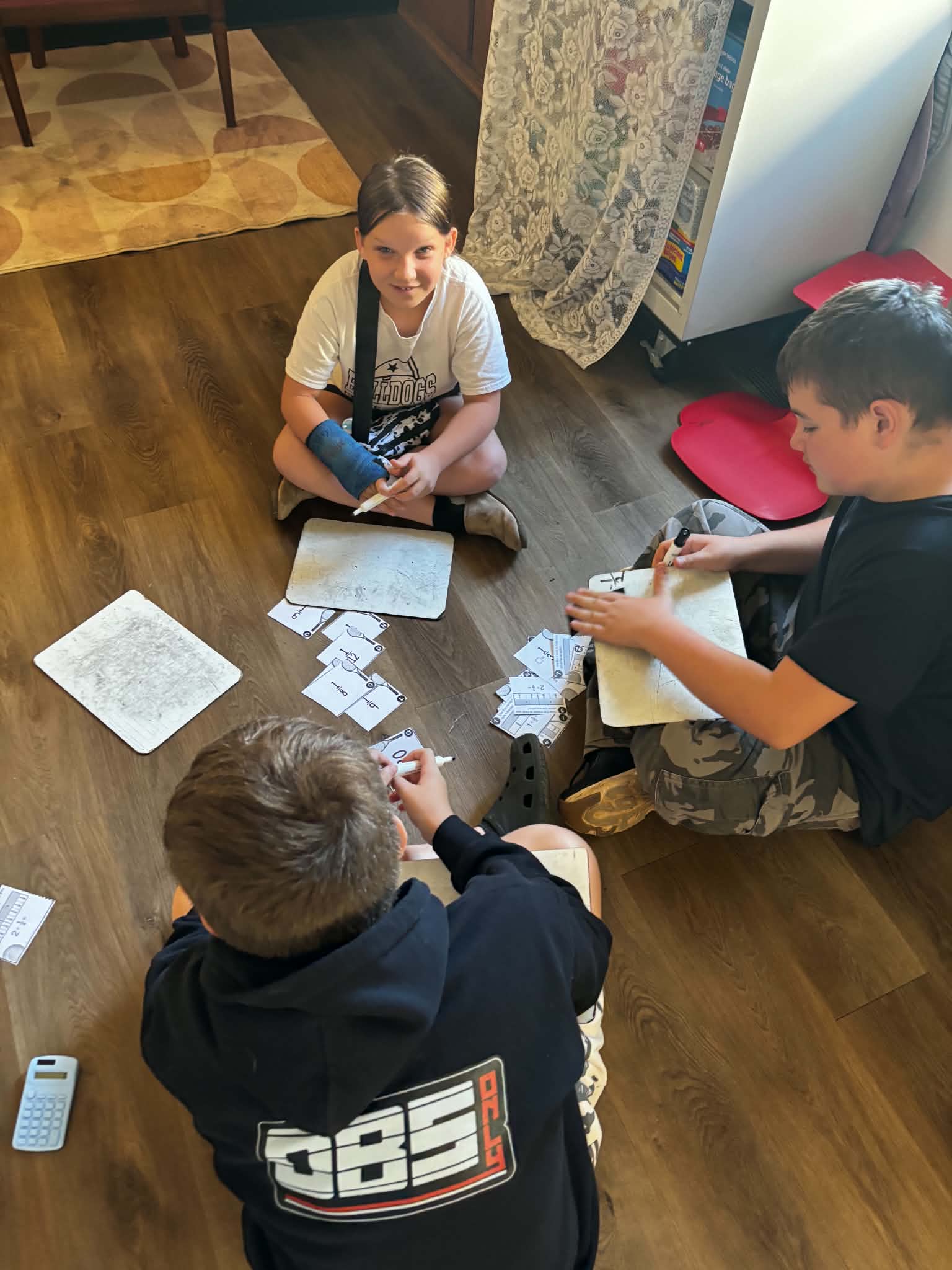 Three children sit on a hardwood floor, working on math flashcards and whiteboards.