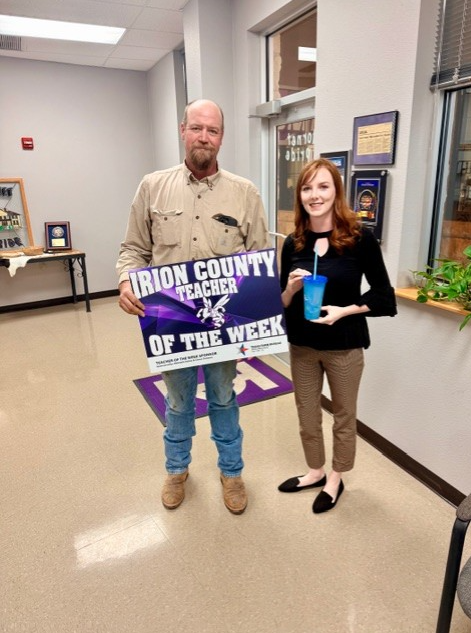 A man holds a sign that reads "IRION COUNTY TEACHER OF THE WEEK" while standing next to a woman holding a blue cup.