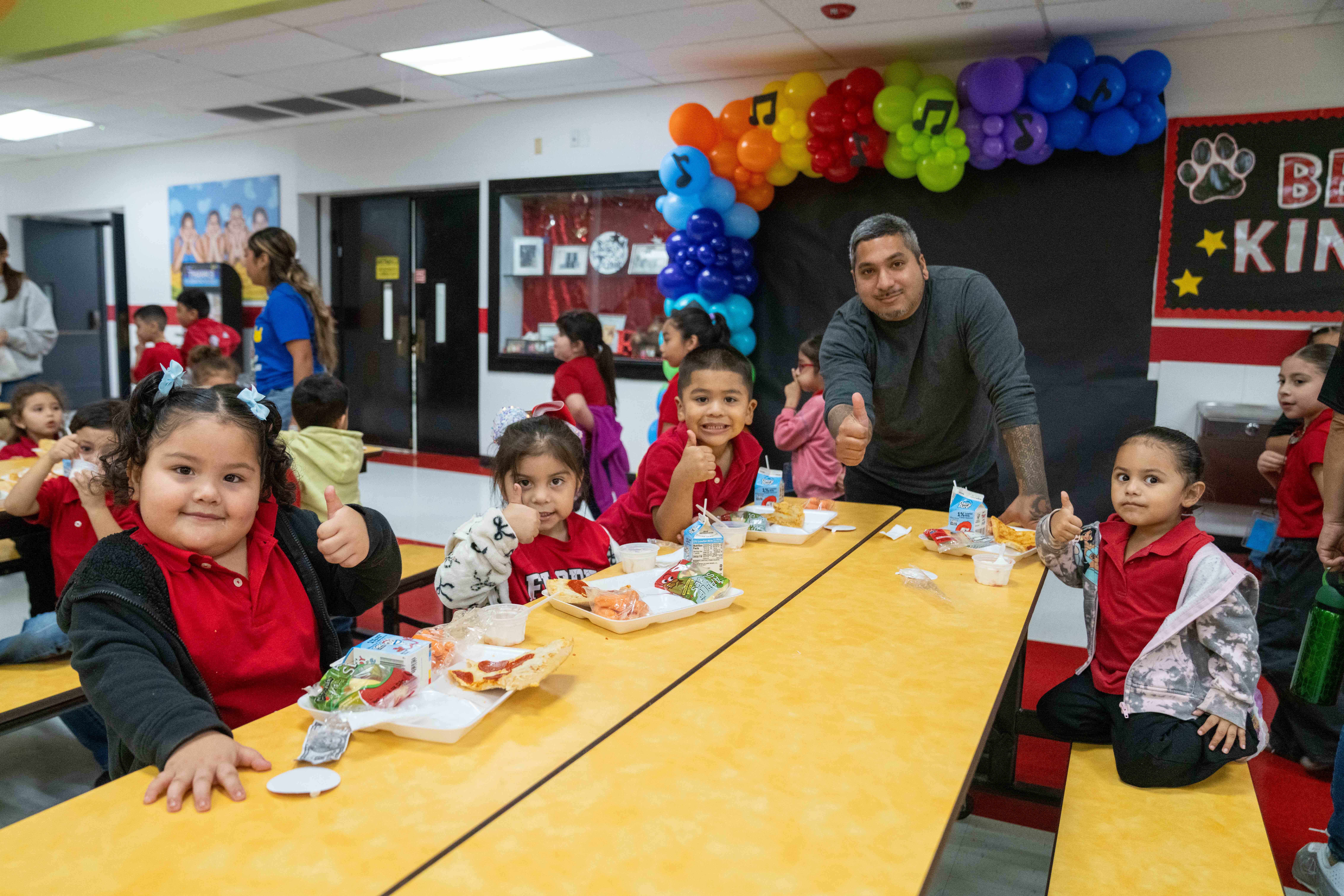 A group of children and an adult give thumbs-up while sitting at lunch tables in a brightly decorated school cafeteria.
