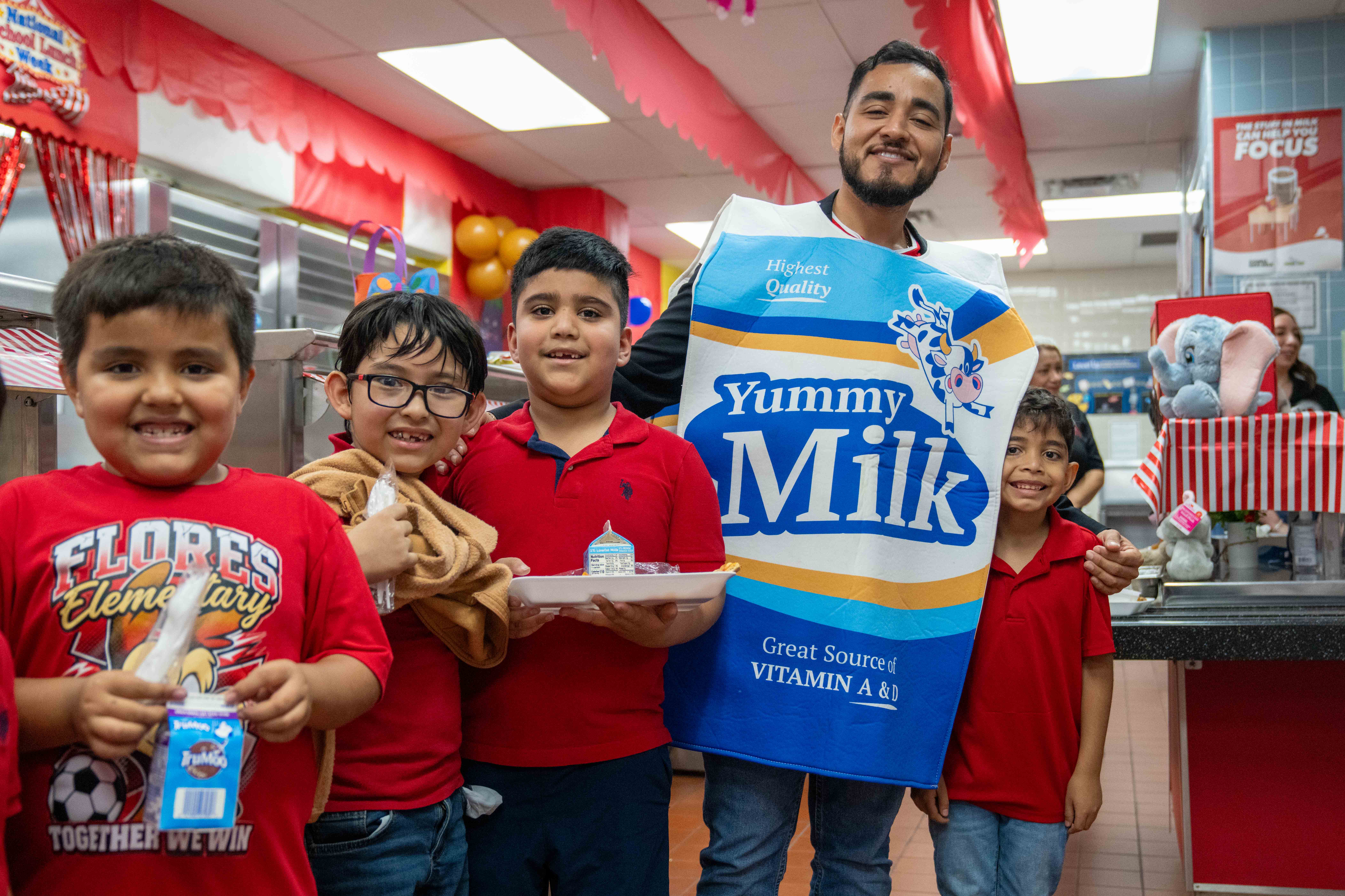 A man in a milk carton costume poses with four smiling children in a school cafeteria.