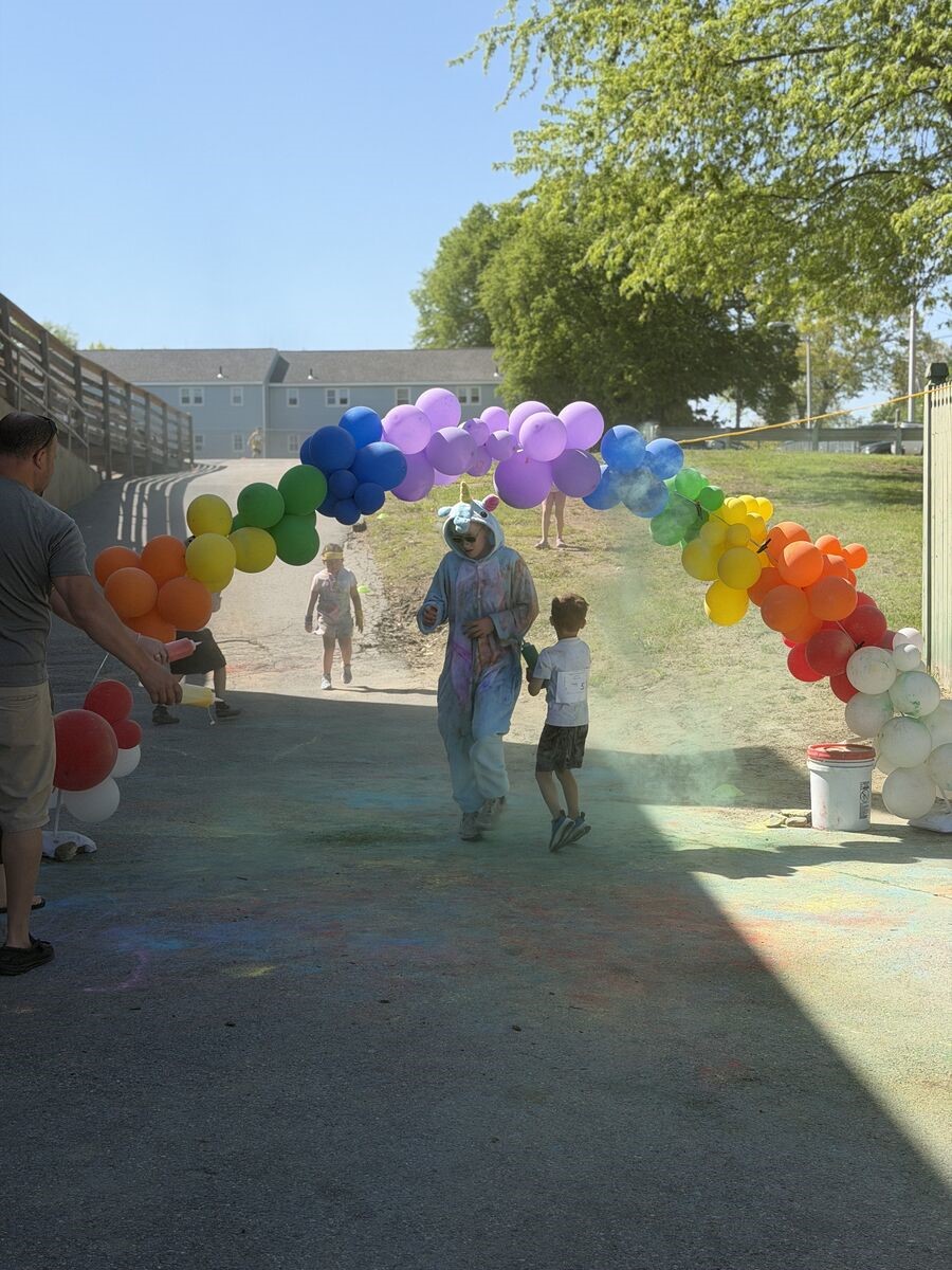 Children run through a rainbow balloon arch at a color run event, with colorful powder on the ground.