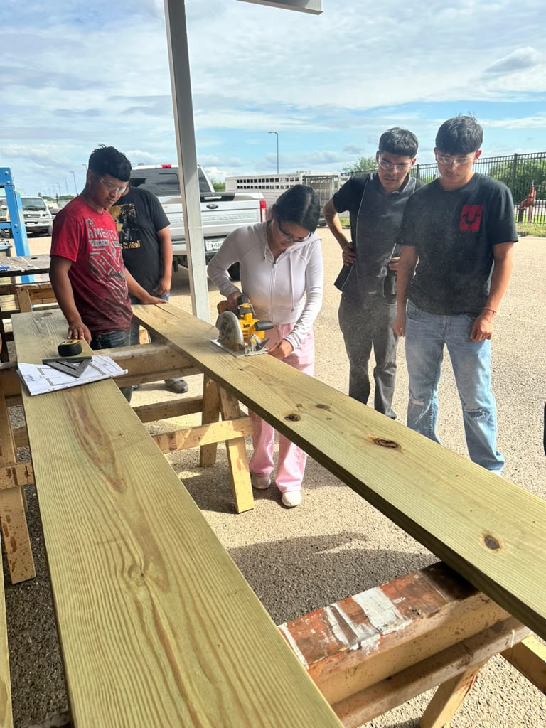 A student uses a circular saw to cut a wooden plank while classmates observe outdoors.