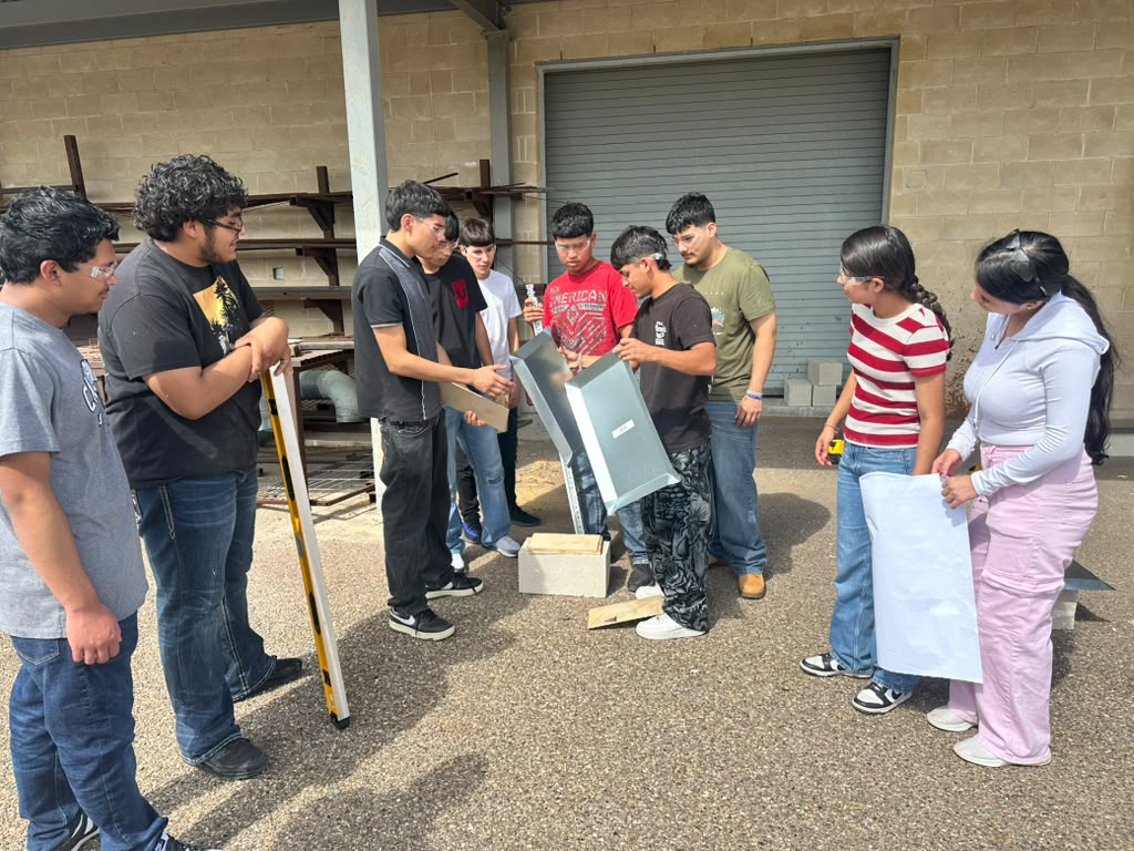 A group of students gather outdoors near a brick building, working together on a metal fabrication project.