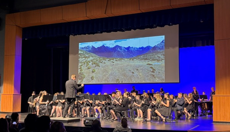A conductor leads a band of musicians on a stage with a scenic mountain landscape projected on a large screen.