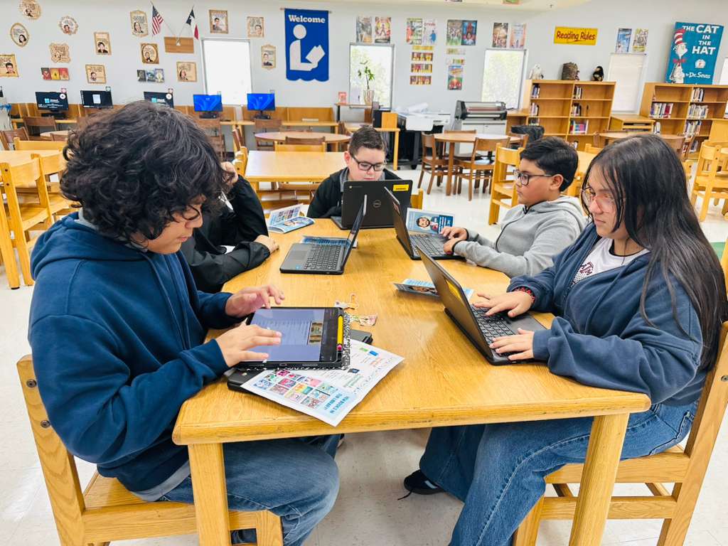 Students work collaboratively on laptops and a tablet at a wooden table in a library.