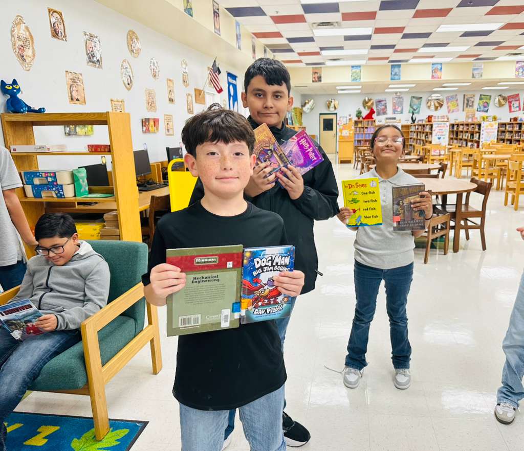Three children proudly hold up books in a library setting, with one child reading in a chair.