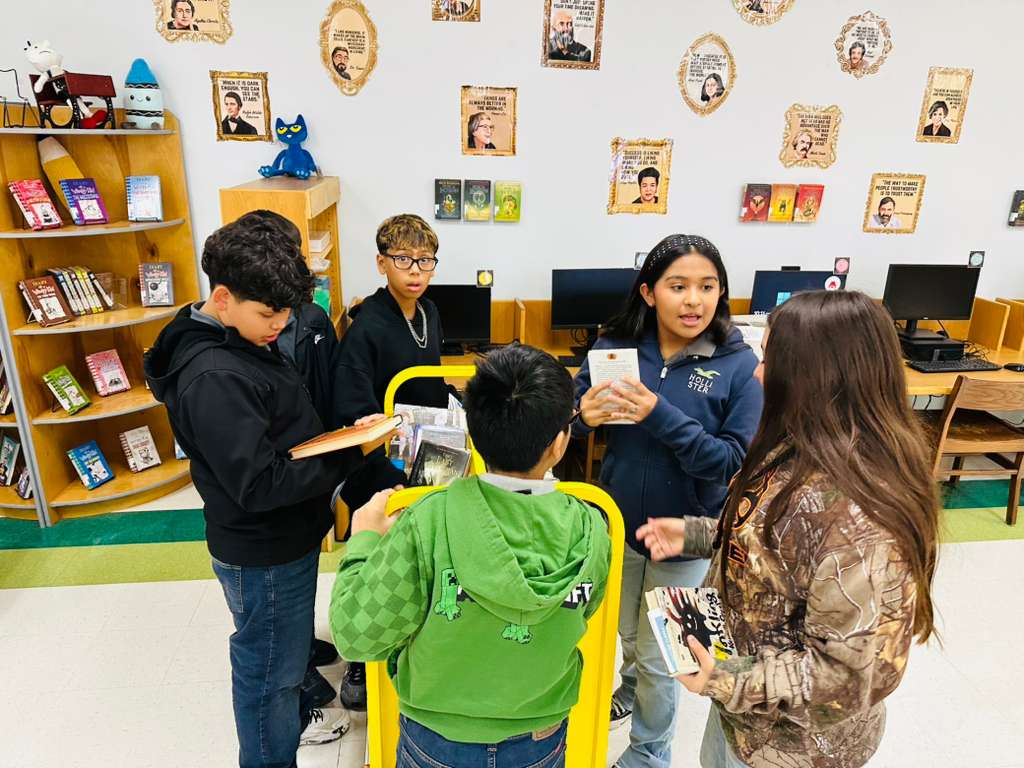 Students gather around a bookshelf in a classroom, discussing books.