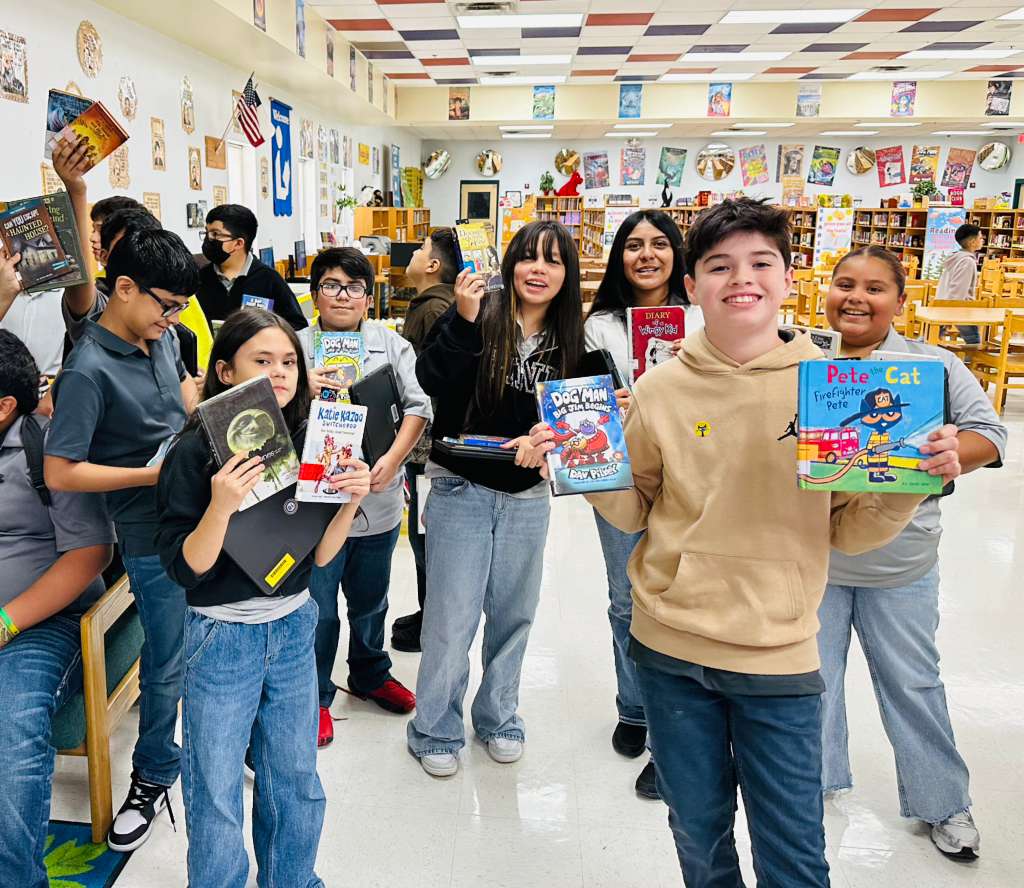 A group of smiling students proudly hold up various books in a library setting.