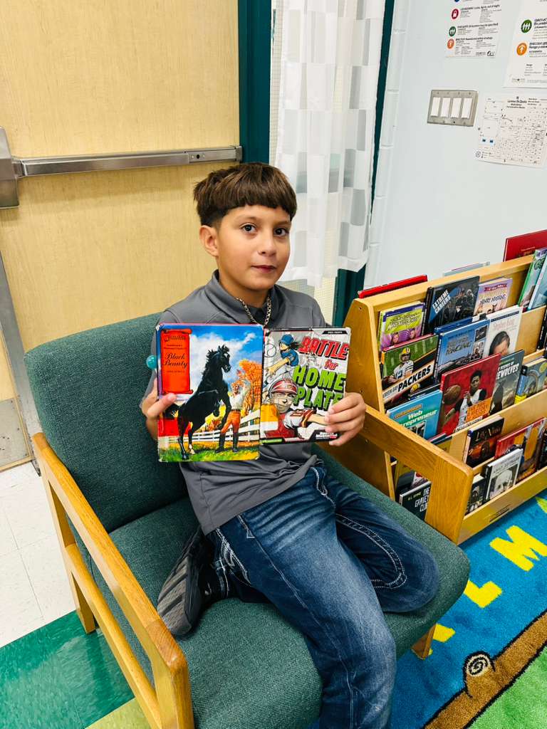 A young boy sits in a chair holding two books, one titled 'Black Beauty' and the other 'Battle Home Plate'.