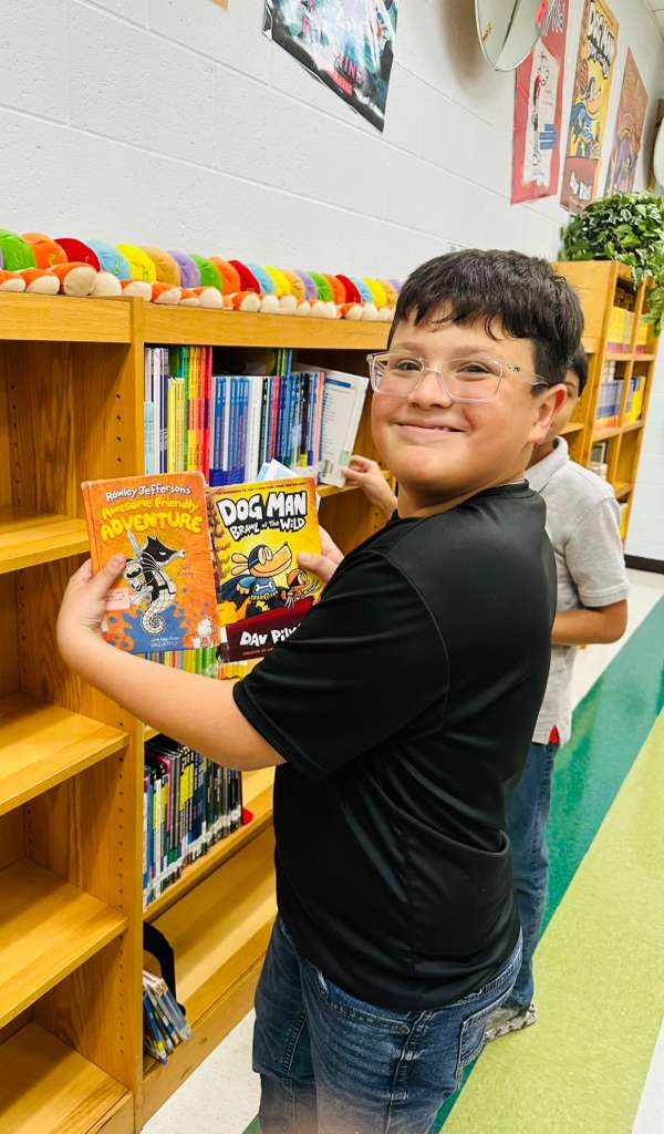 A young boy with glasses smiles while holding two books, 'Awesome Friendly Adventure' and 'Dog Man: Brawl of the Wild', in a library.