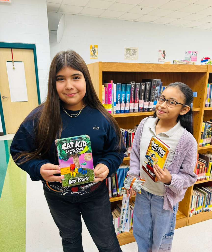 Two young girls stand in a library, each holding a book by Dav Pilkey.