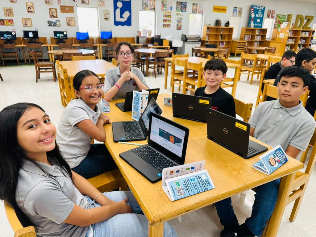 Students work on laptops at tables in a bright, modern library setting.