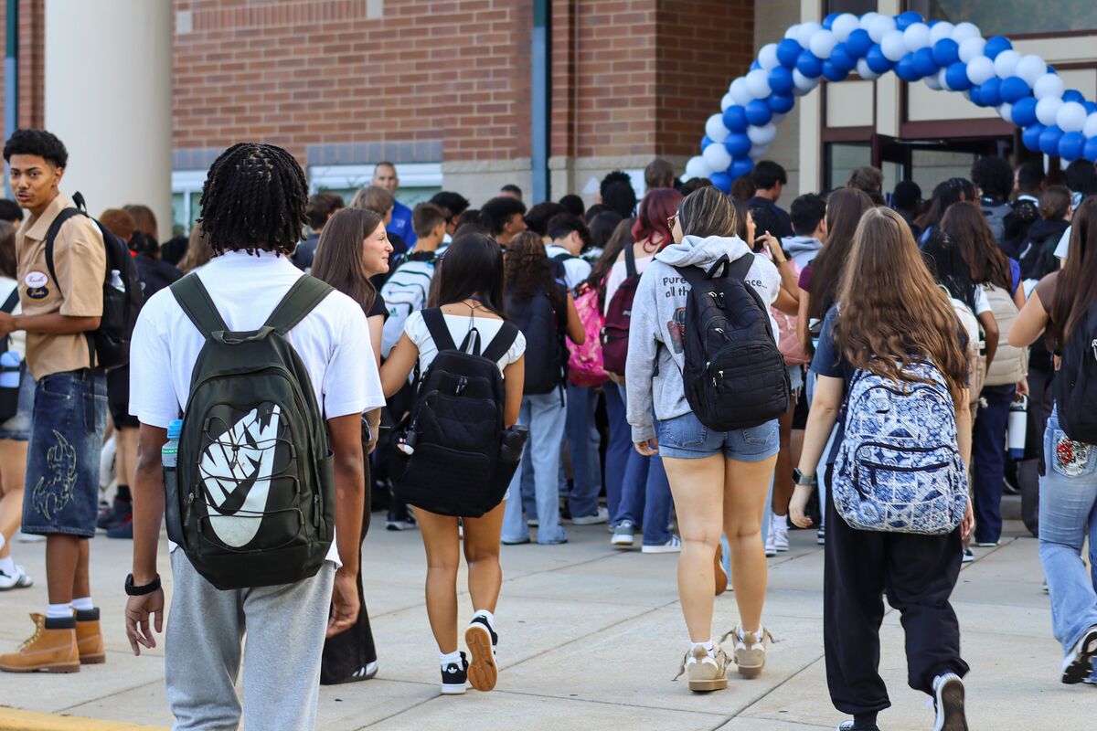 A diverse group of students with backpacks walks together toward a school entrance decorated with a blue and white balloon arch.
