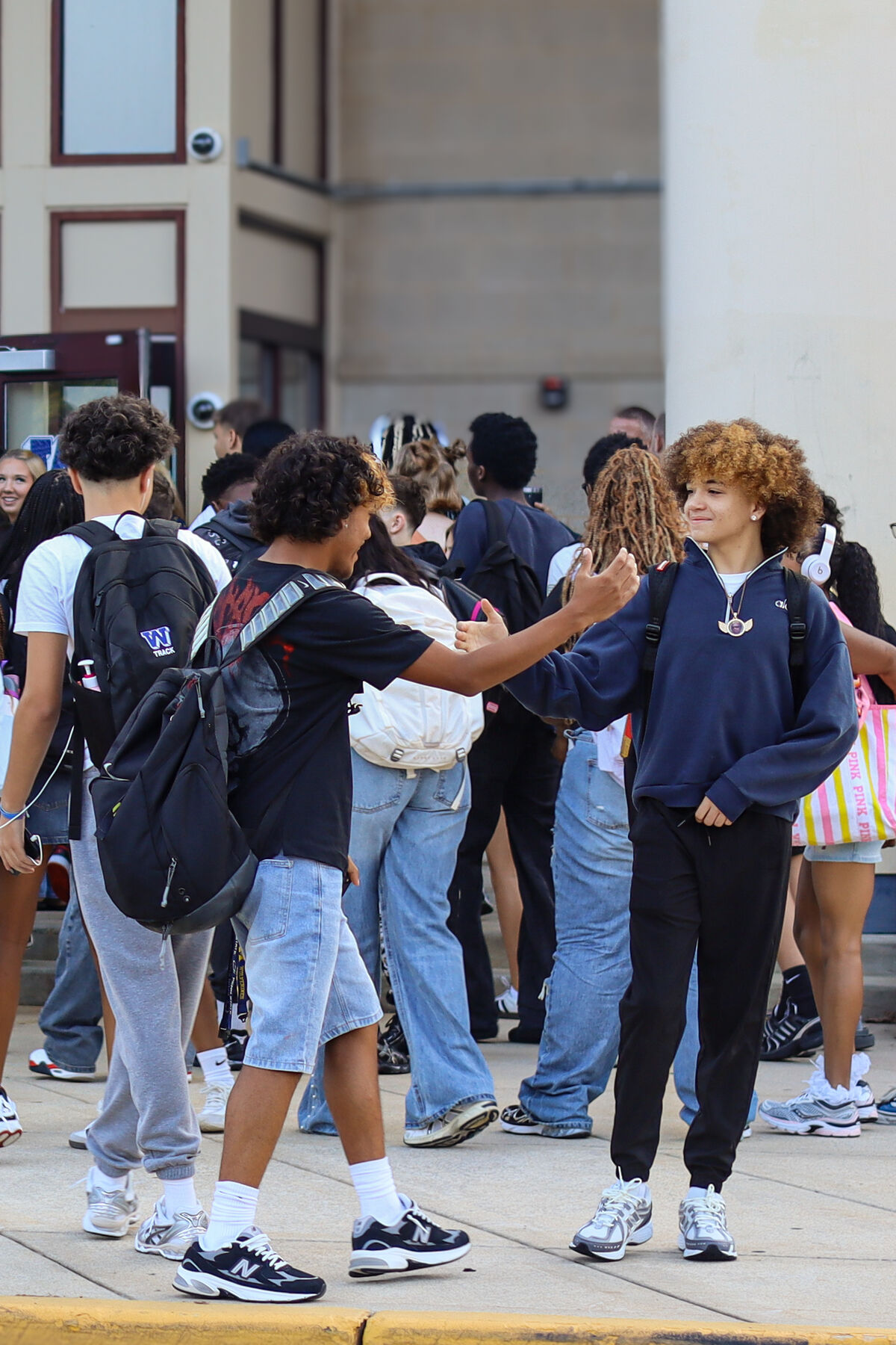 Two teenagers greet each other outdoors, surrounded by a crowd of students near a building entrance.