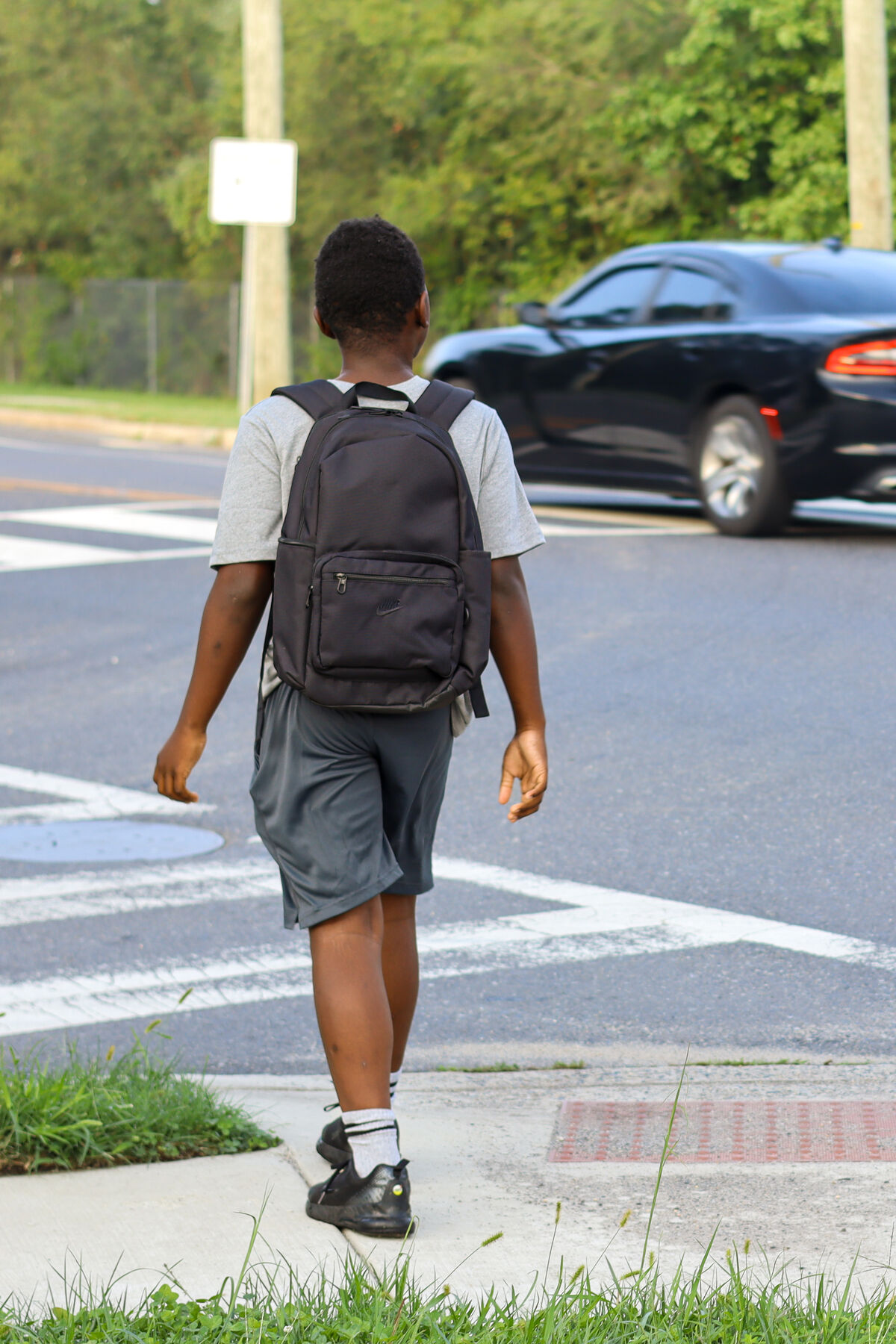 A young person with a black backpack walks along a sidewalk near a crosswalk.