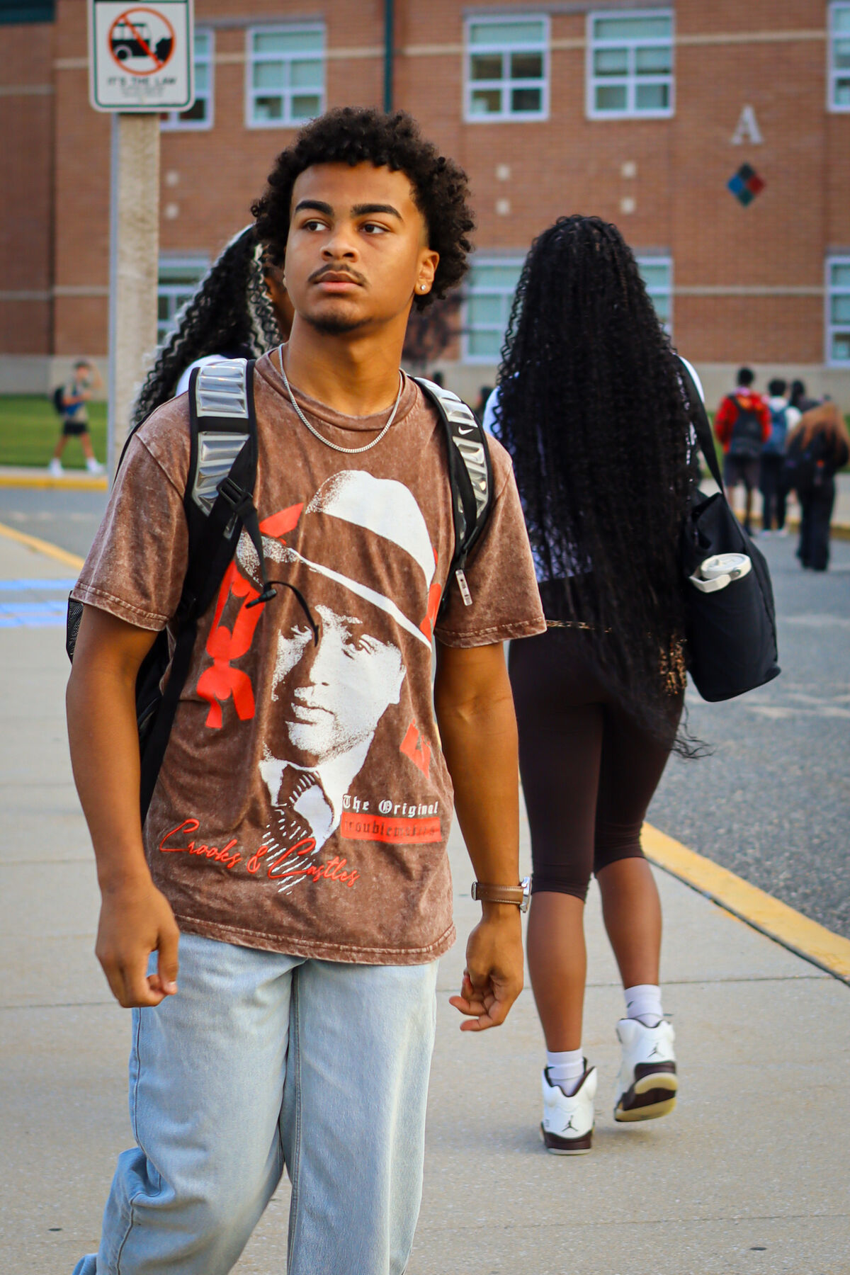 A young man with curly hair and a graphic t-shirt walks outdoors near a brick building.