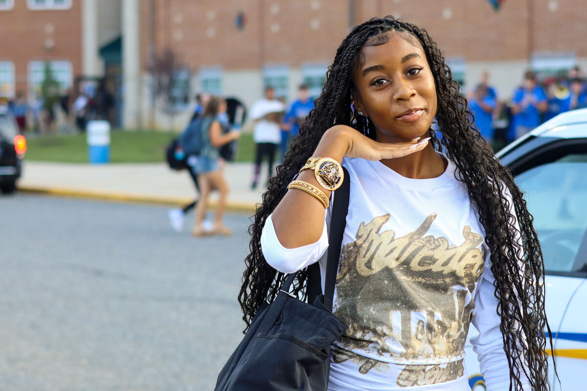 A young person with long braided hair poses outdoors, wearing a white shirt and gold bracelets.