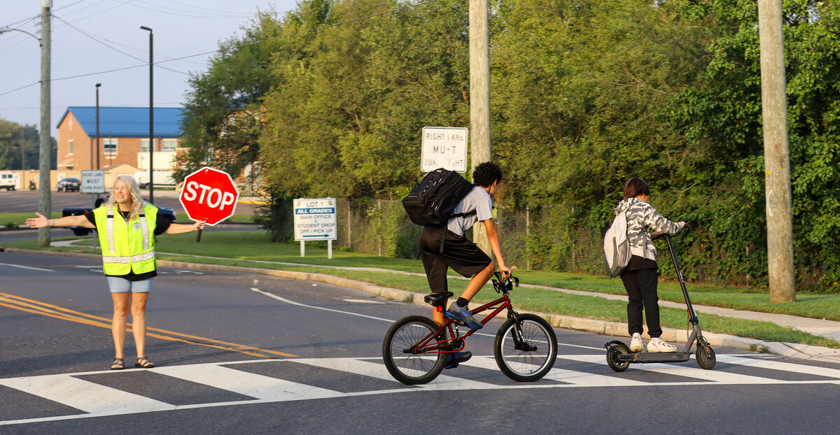 A crossing guard in a high-visibility vest holds a stop sign as two students cross the street.