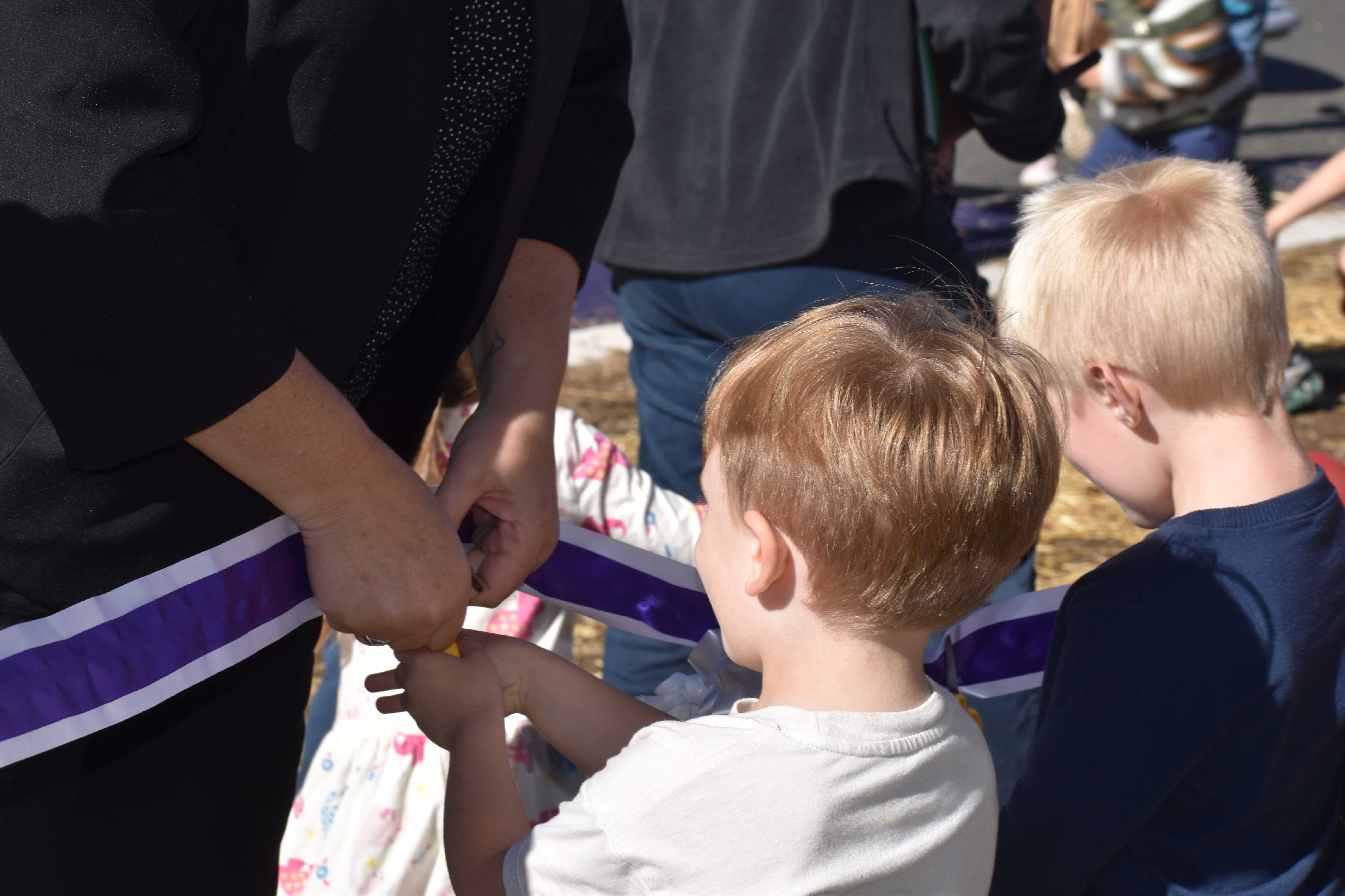 A child in a white shirt holds a purple ribbon alongside an adult.