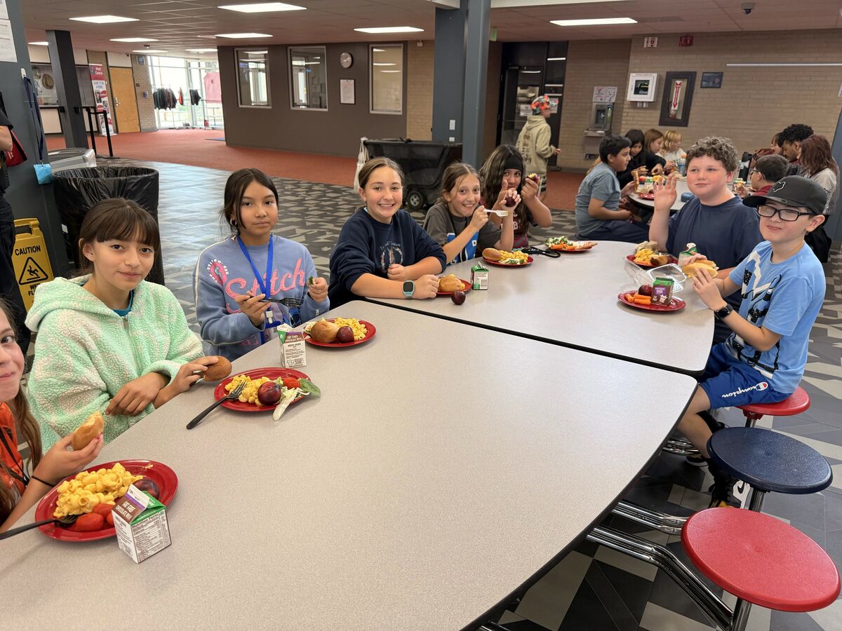 A group of students sits at long cafeteria tables, eating lunch and smiling for the camera.