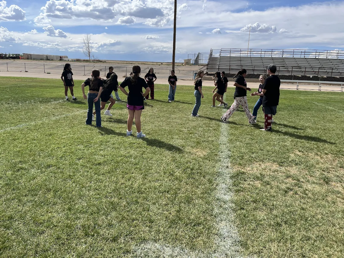 A group of students stands on a grassy athletic field under a partly cloudy sky.