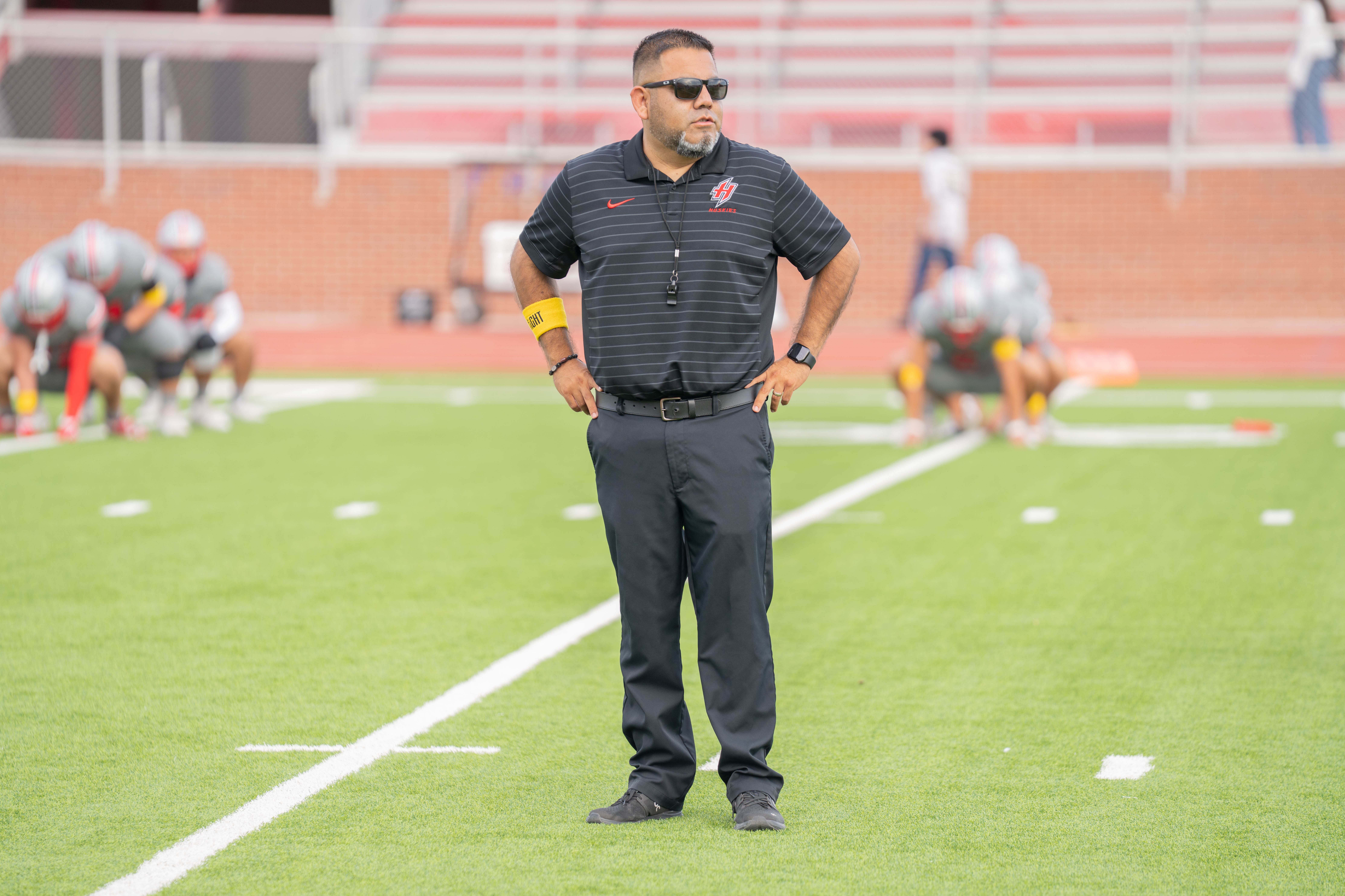 A coach stands on a football field with hands on hips, watching players practice in the background.