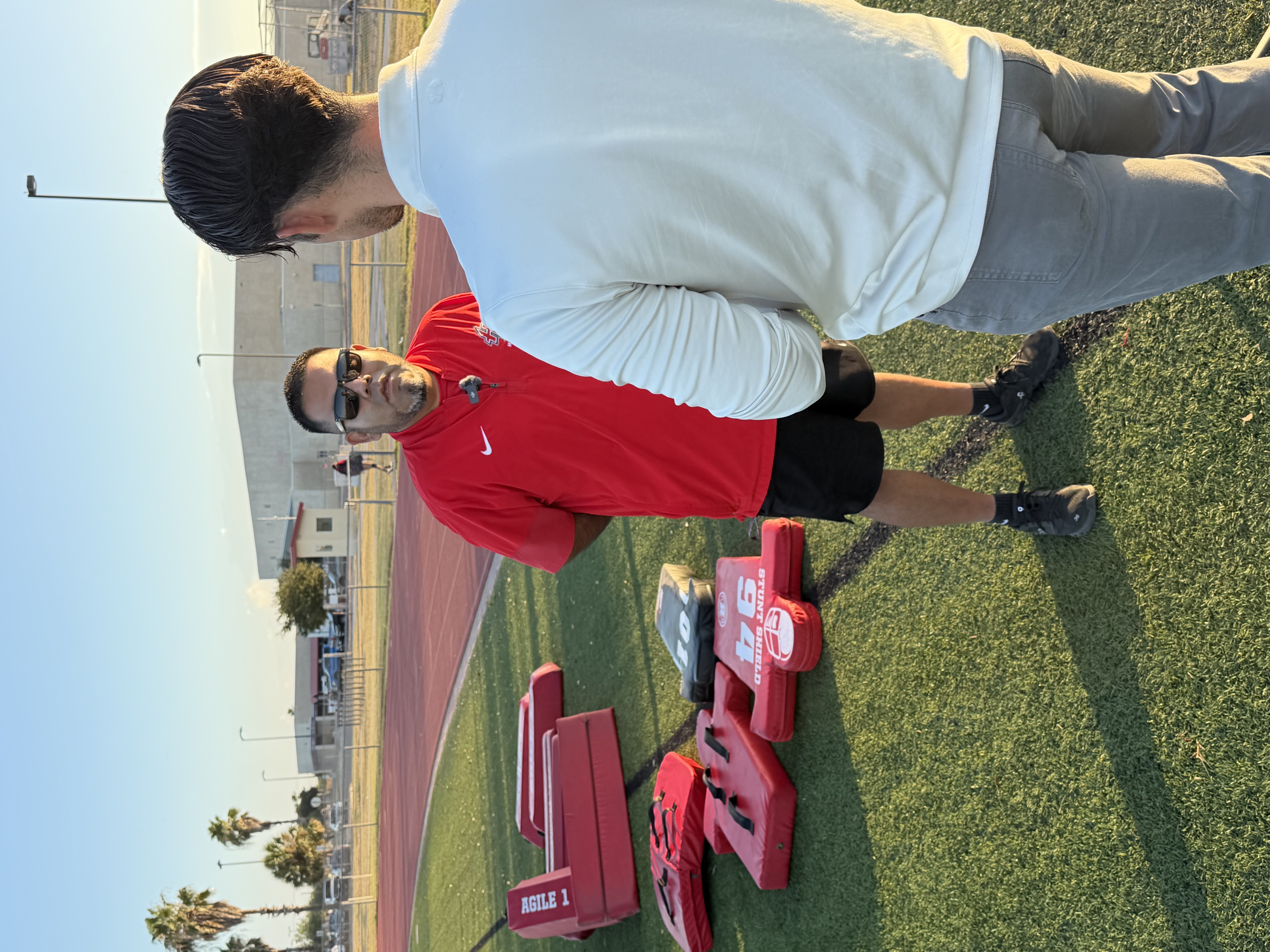 Two men stand outside on a turf field next to red athletic training equipment.